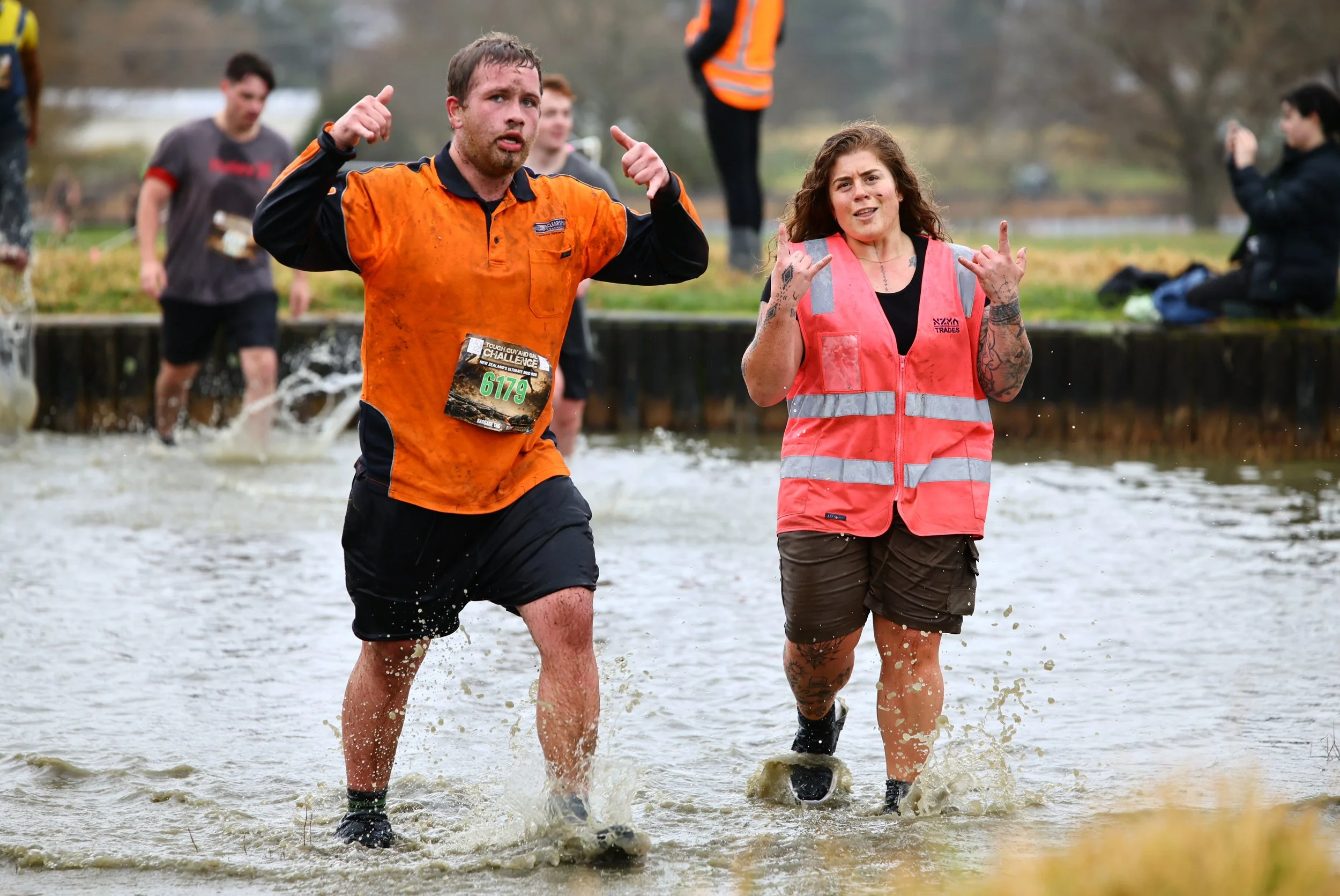 People running through a muddy water obstacle course on a rainy day, with some participants wearing numbered bibs and a person in a pink safety vest in the foreground.