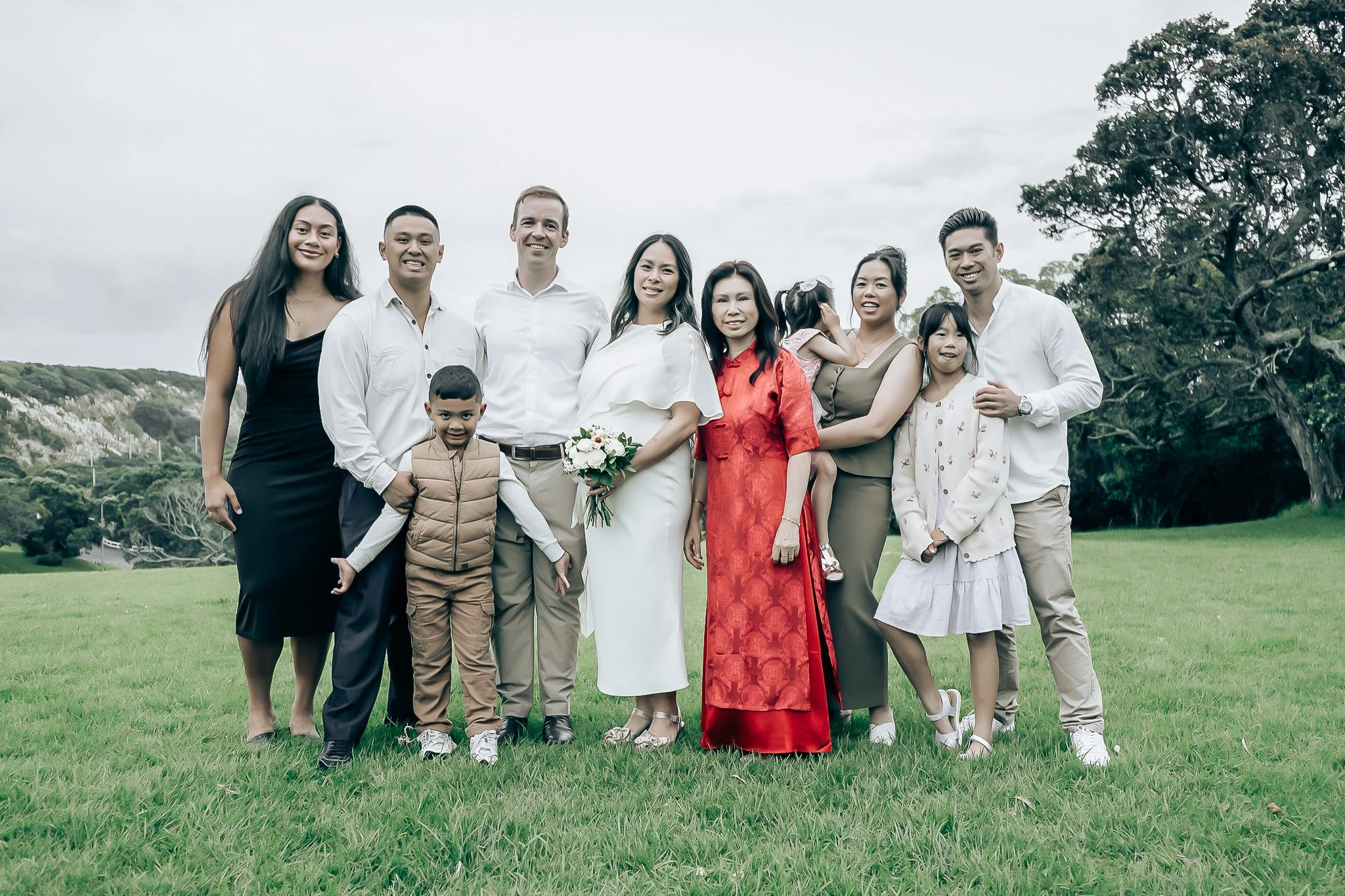 A group of ten diverse people, including children, standing together on a grassy field with trees and hills in the background, smiling at the camera.