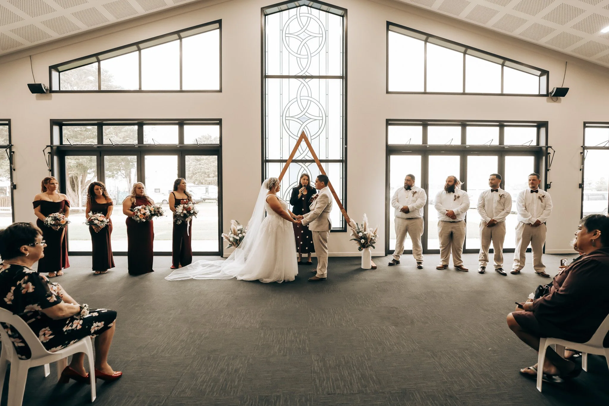 A wedding ceremony taking place indoors with a bride and groom exchanging vows, surrounded by bridesmaids and groomsmen, with guests seated and watching.