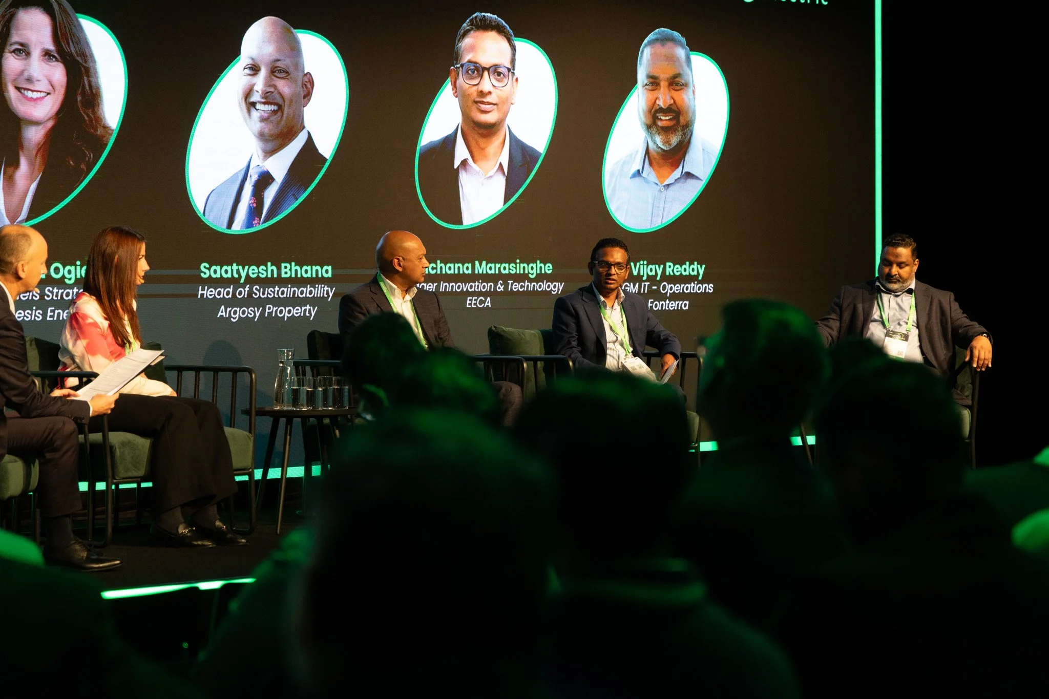 Panel of professionals sitting on a stage during a conference, with a large digital screen behind them displaying headshots and names of speakers.
