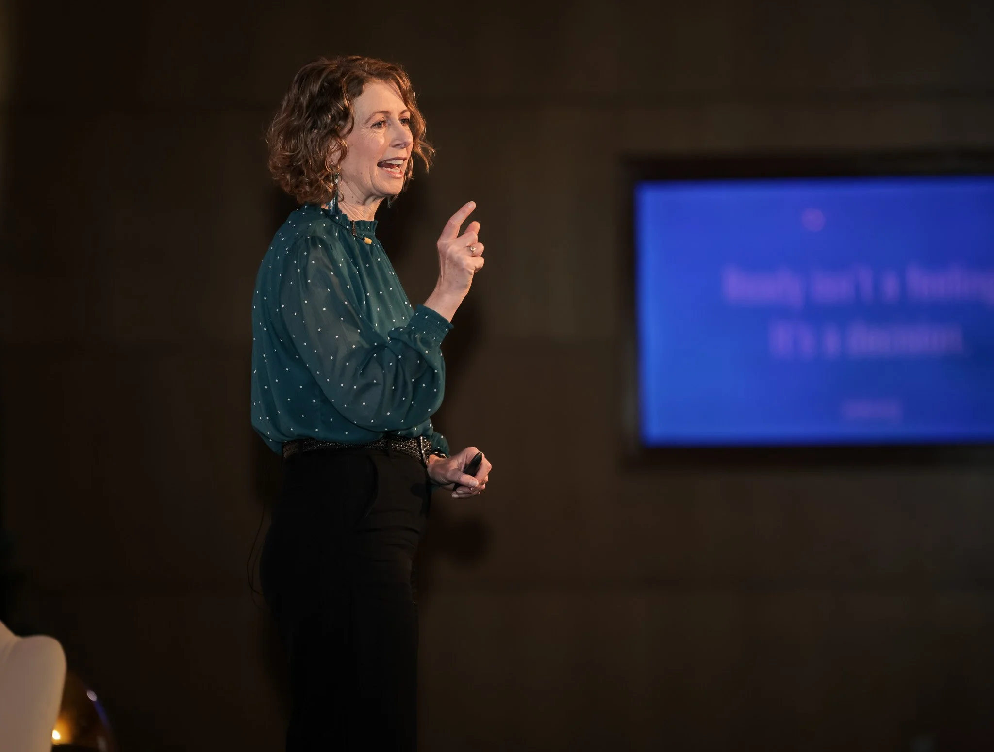 A woman with curly hair wearing a dark green blouse with white polka dots giving a presentation inside a conference room.