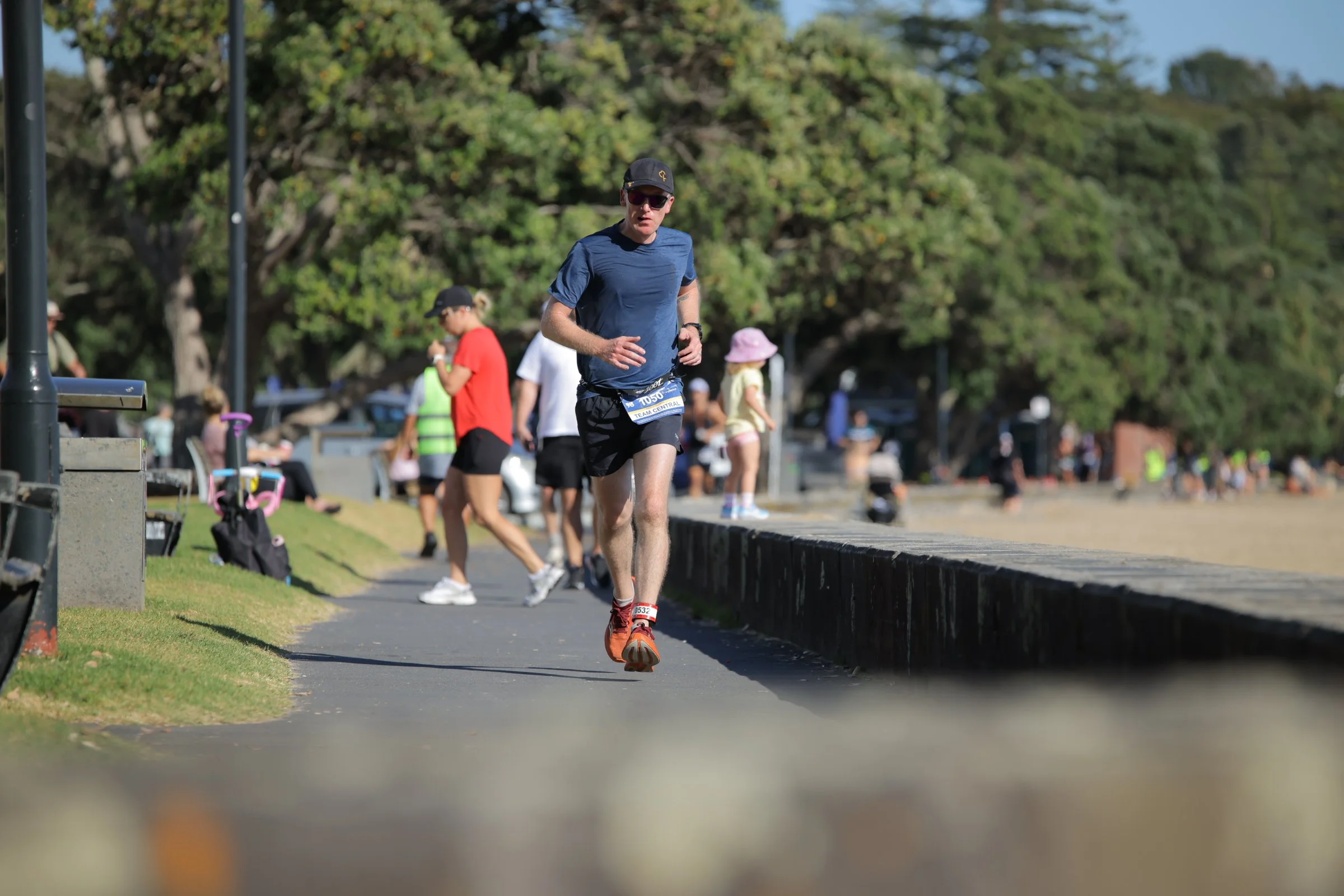 A man running in a race or marathon event, wearing a blue T-shirt, black shorts, orange running shoes, a black cap, and sunglasses, with a race bib number T505. The background includes other participants, some standing and some walking, a green leafy