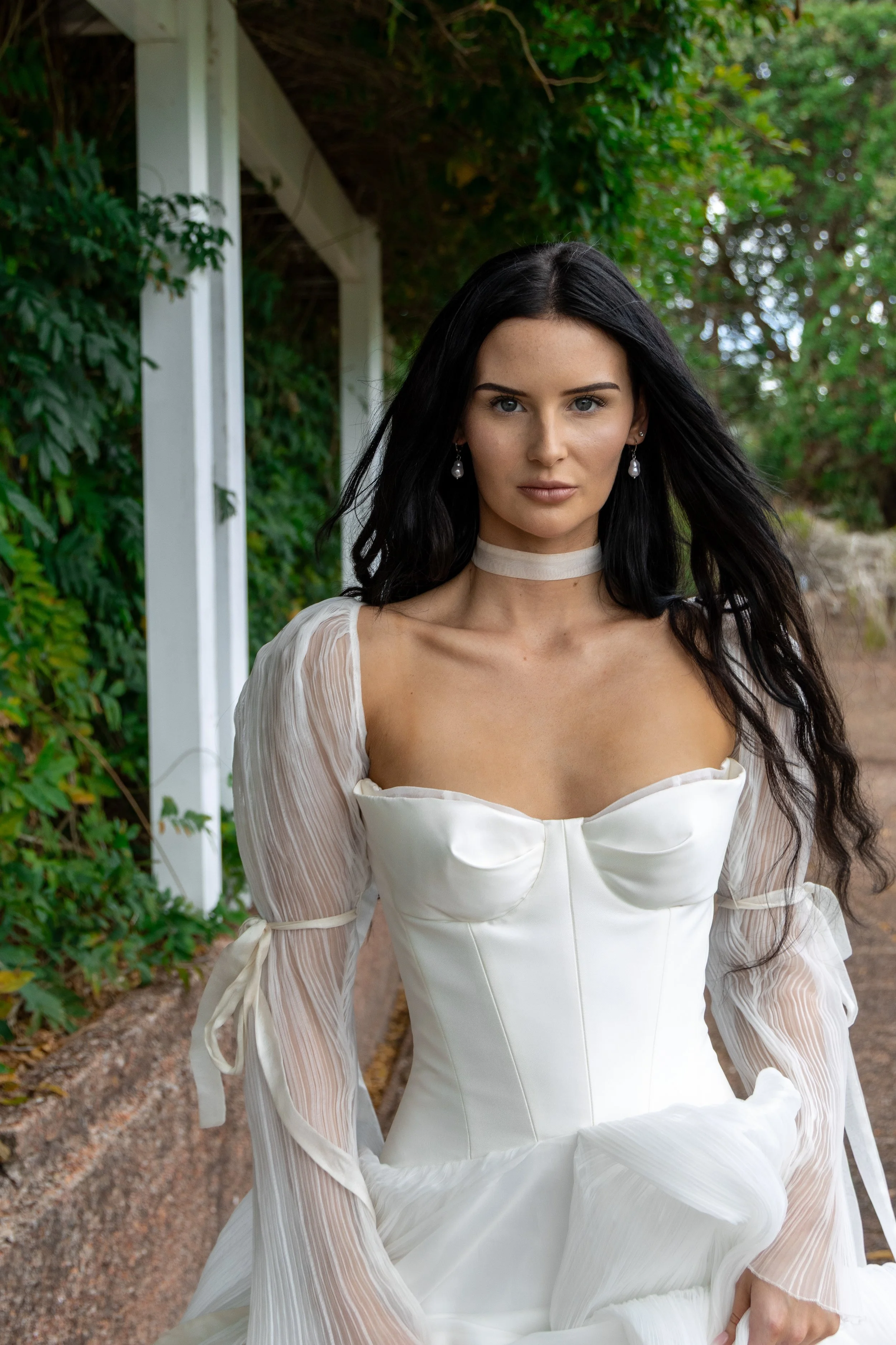 A woman with long black hair, wearing pearl earrings, a cream choker, and a white dress with sheer long sleeves and a bow detail on the chest, standing outdoors in front of green foliage and a white structure.