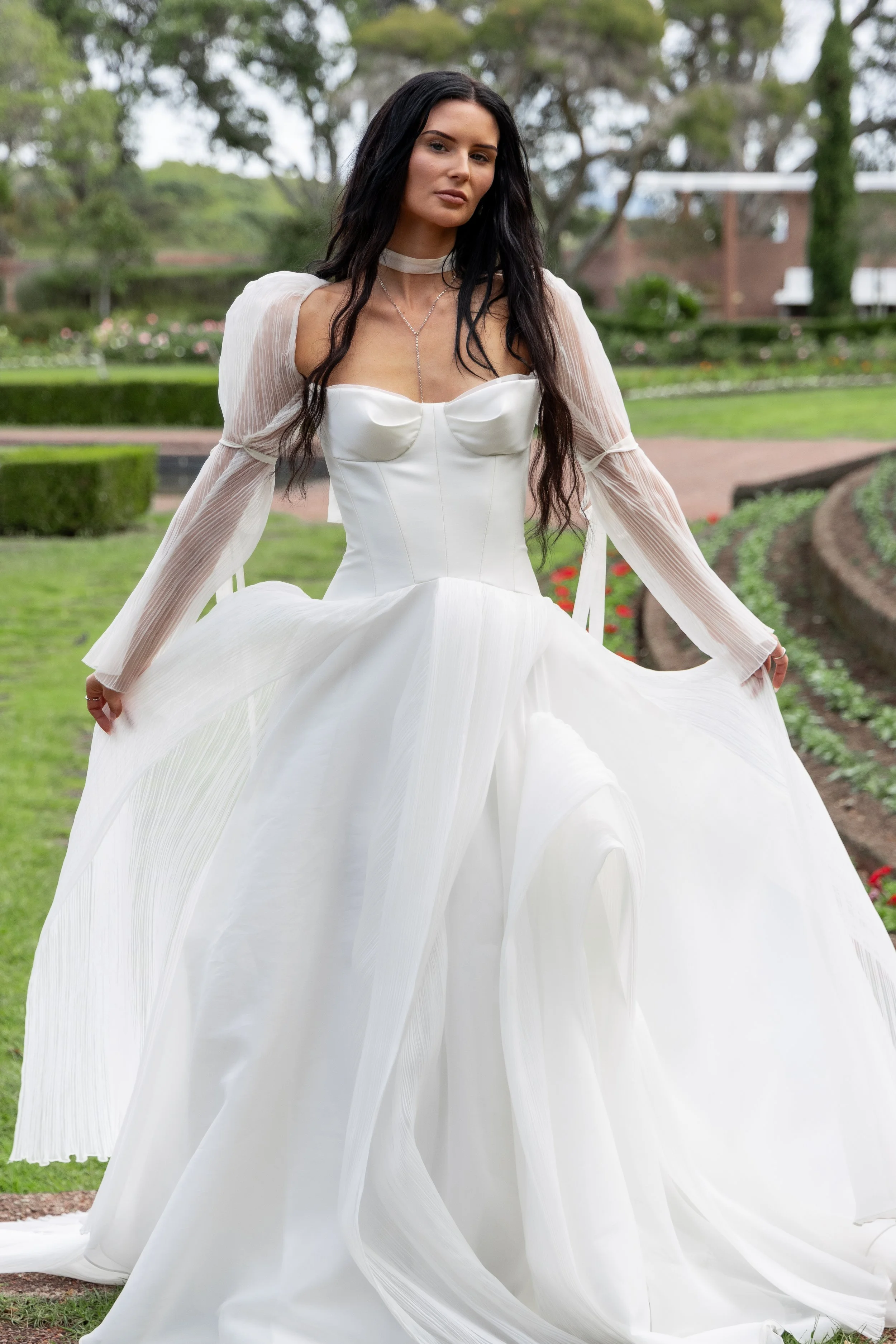 A woman with long dark hair wearing a white wedding dress outdoors in a park with green trees and bushes in the background.