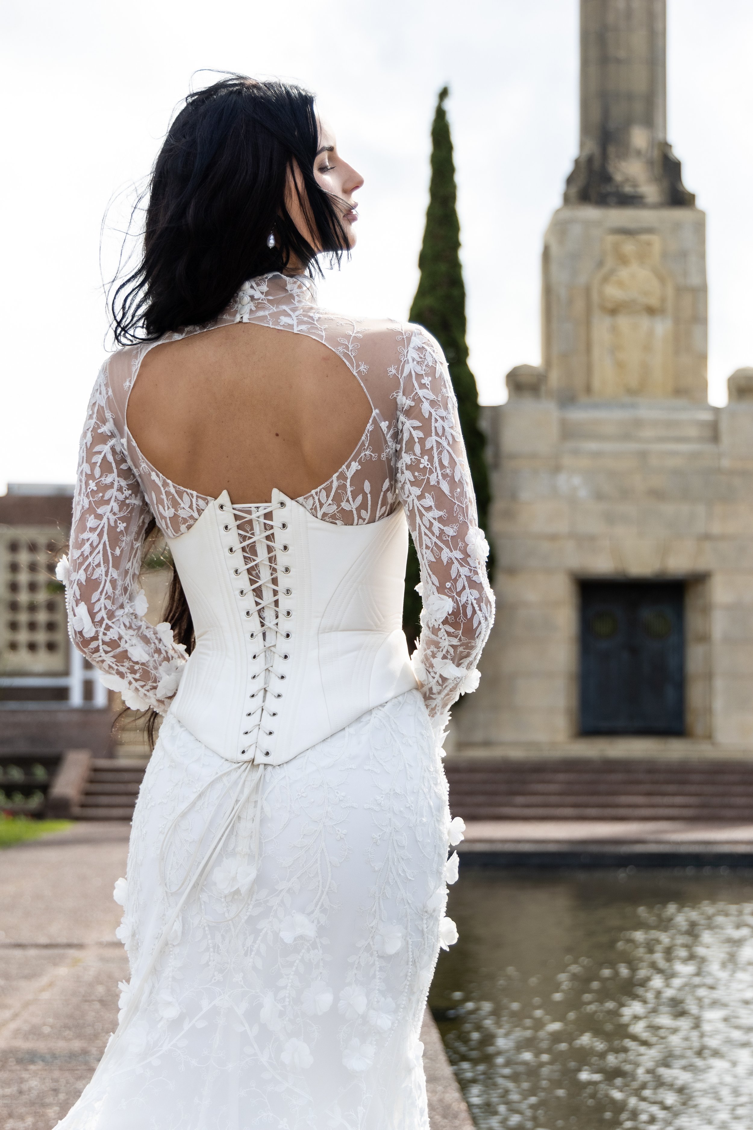 A woman with dark hair and pearl earrings wearing a white lace wedding dress standing by a river with stone steps, a monument, and tall trees in the background.