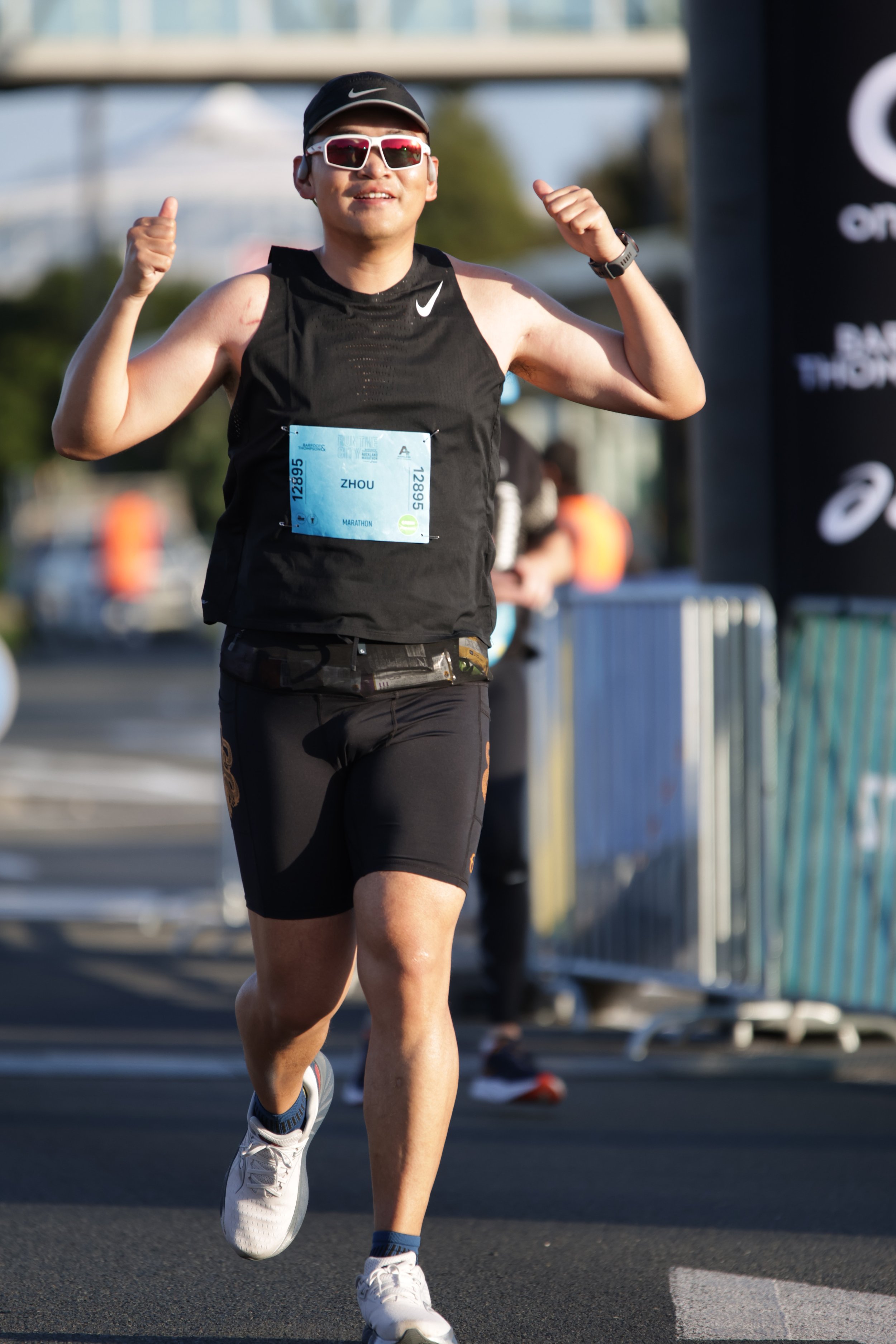 A young man in athletic gear running outdoors during a marathon, wearing sunglasses, a black Nike tank top, and a race bib numbered 12895.