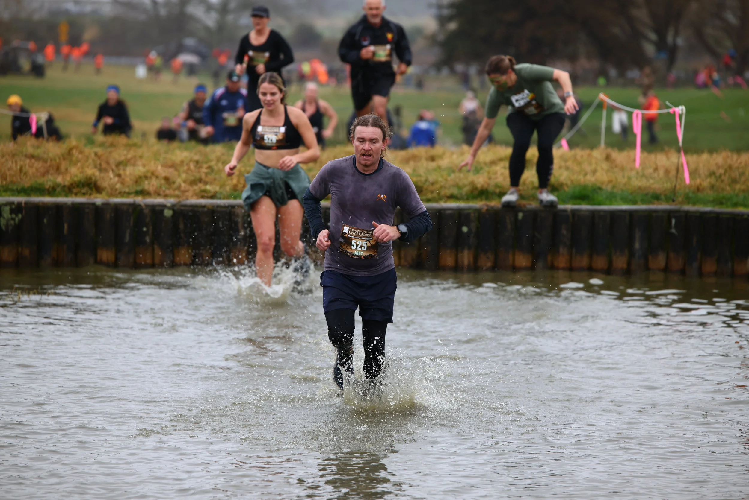A man running through a water obstacle during an outdoor cross-country race, with other runners and spectators in the background.