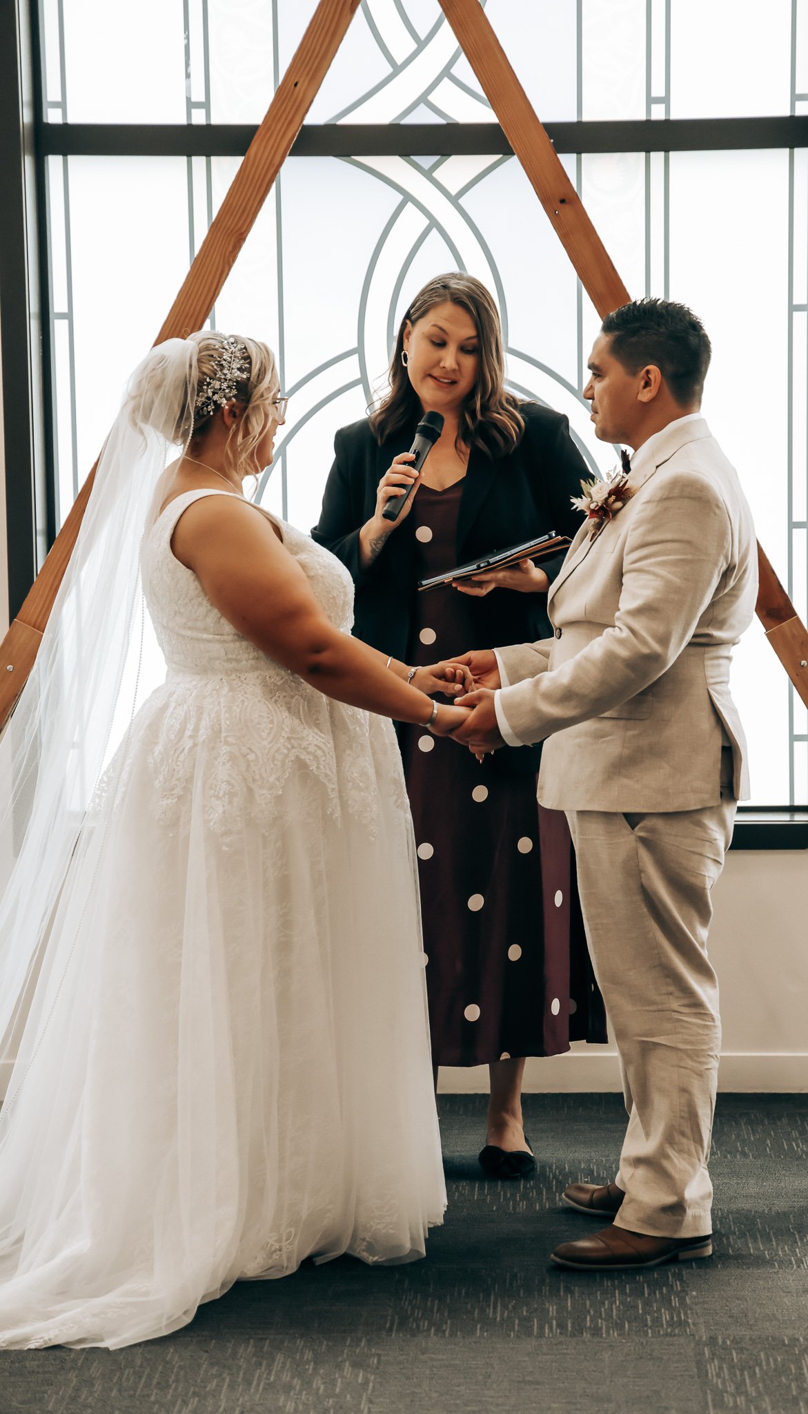 A couple getting married, holding hands and exchanging vows, standing in front of officiant during wedding ceremony in front of a decorative window.