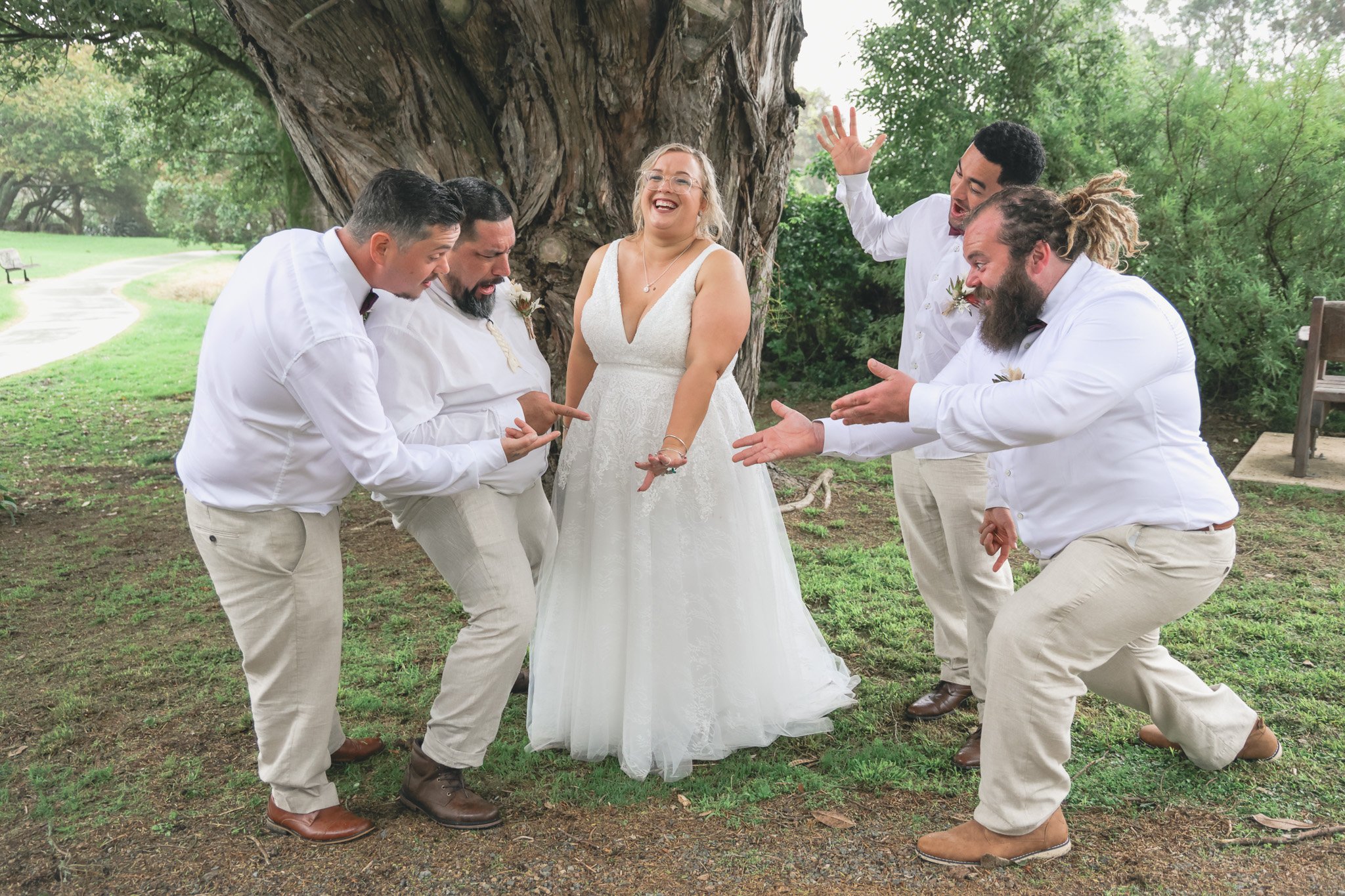 A group of five people, four men and one woman, wearing white shirts and beige pants, are gathered outdoors in front of a large tree. They are smiling, laughing, and engaged in a playful moment. The woman, dressed in a white dress, is at the center w
