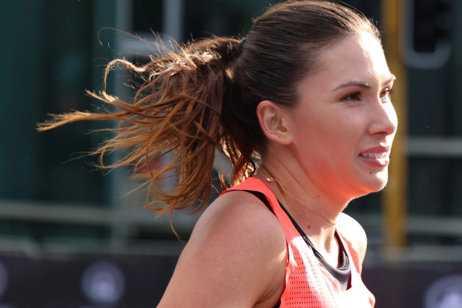 A female athlete with brown hair in a ponytail, wearing a red sports top, running outdoors with a determined expression.