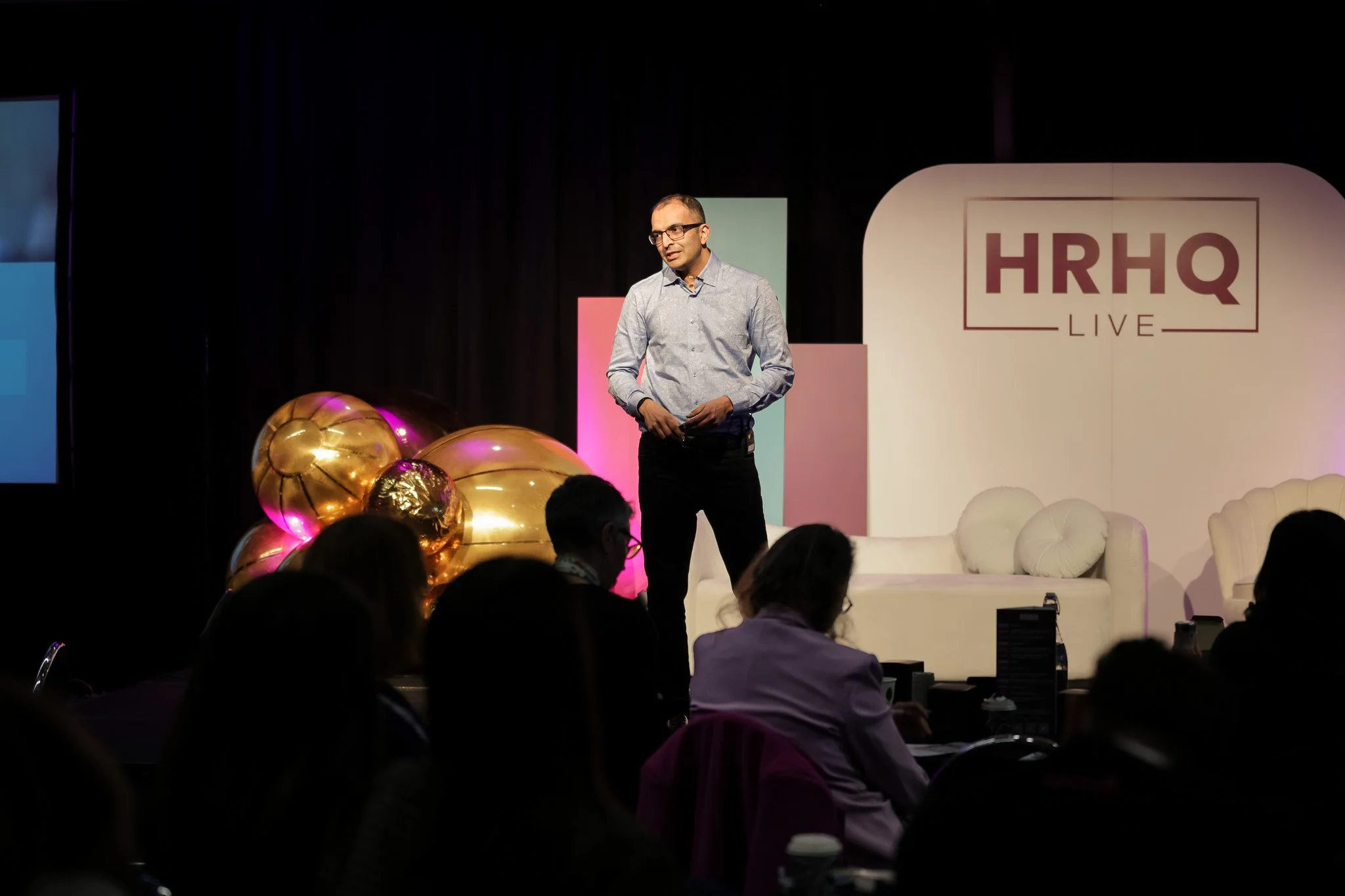 A man speaking on stage at a conference with a backdrop displaying 'HRHQ LIVE'; gold and purple balloons are on the stage, and audience members are seated in front.