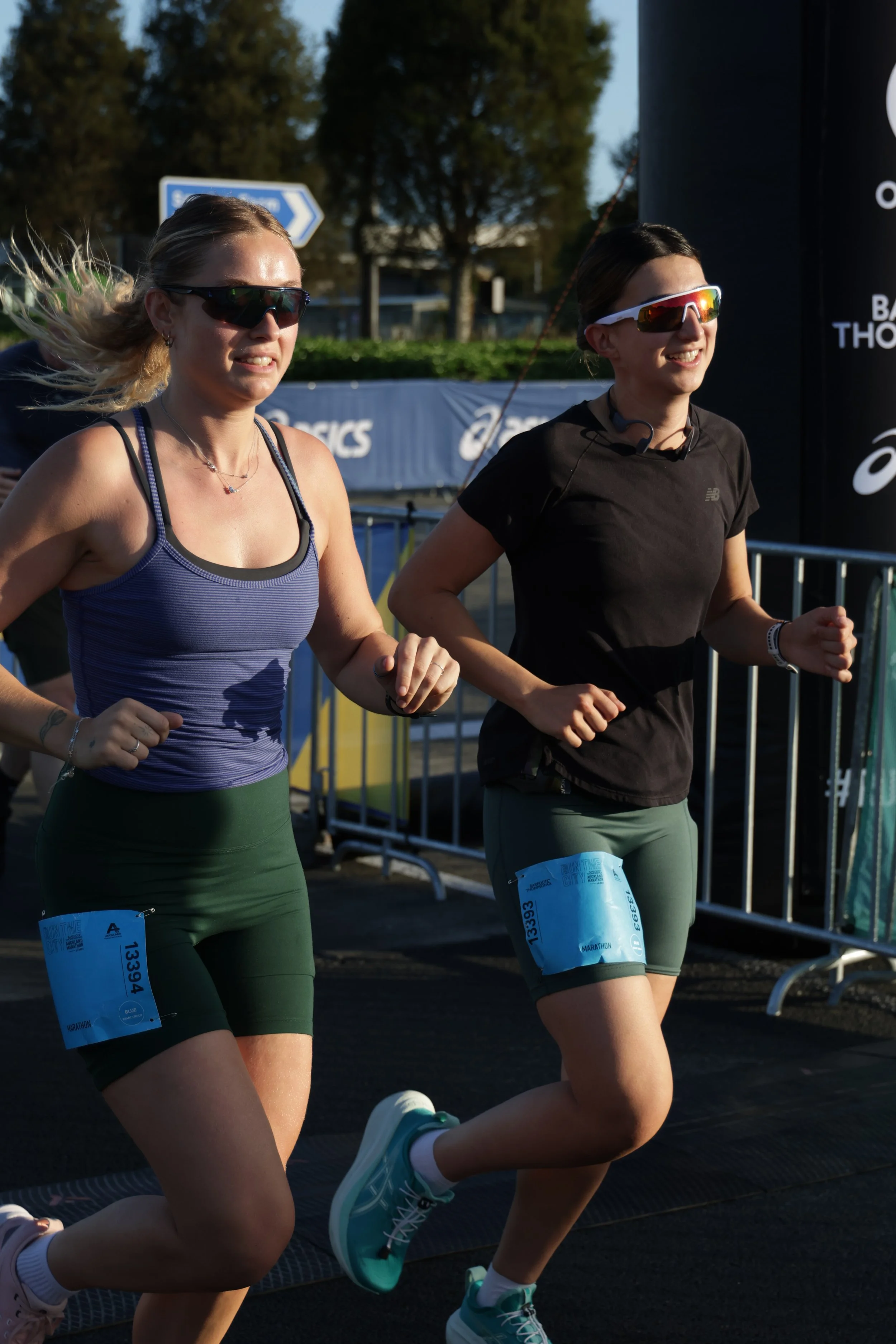 Two female runners participating in a marathon, running outdoors during a sunny day, with race bibs attached to their green shorts, wearing athletic sunglasses and running gear.