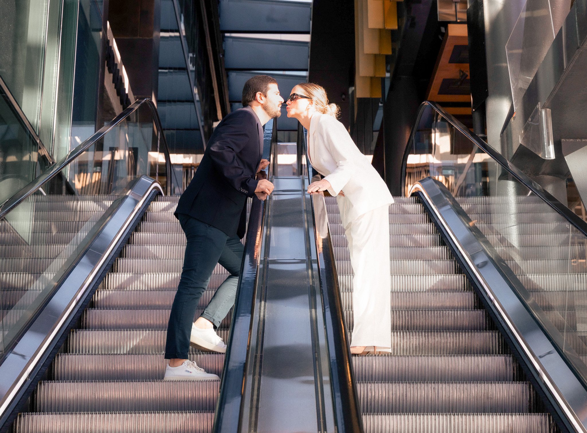 A man and woman standing on escalators in a modern building, leaning toward each other ready to kiss.