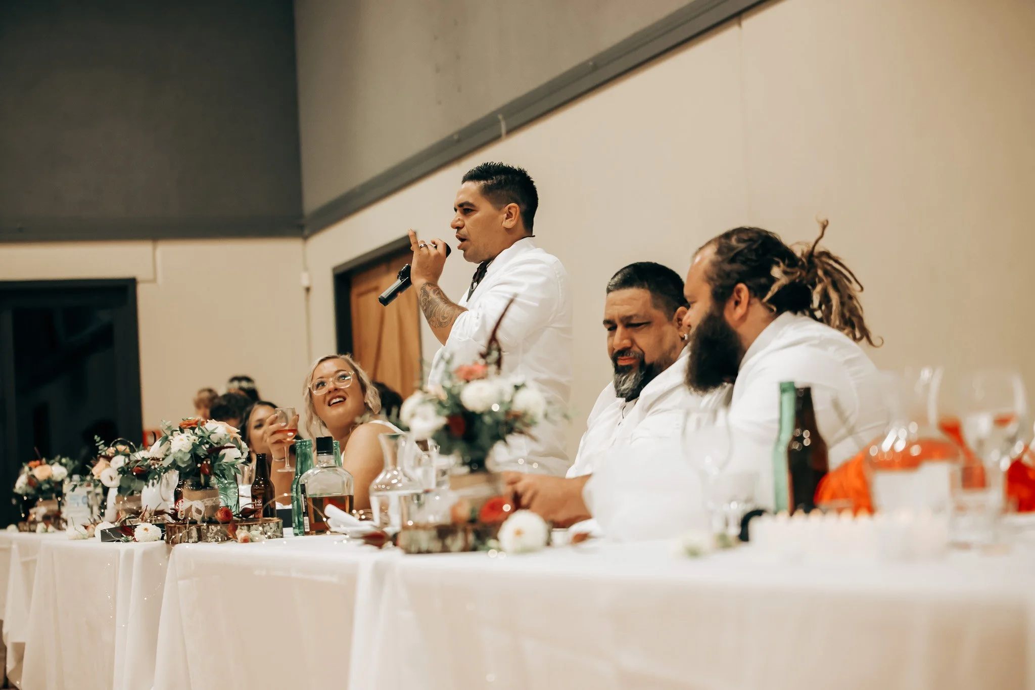 People sitting at a banquet table decorated with flowers and food, with a man standing and speaking into a microphone.