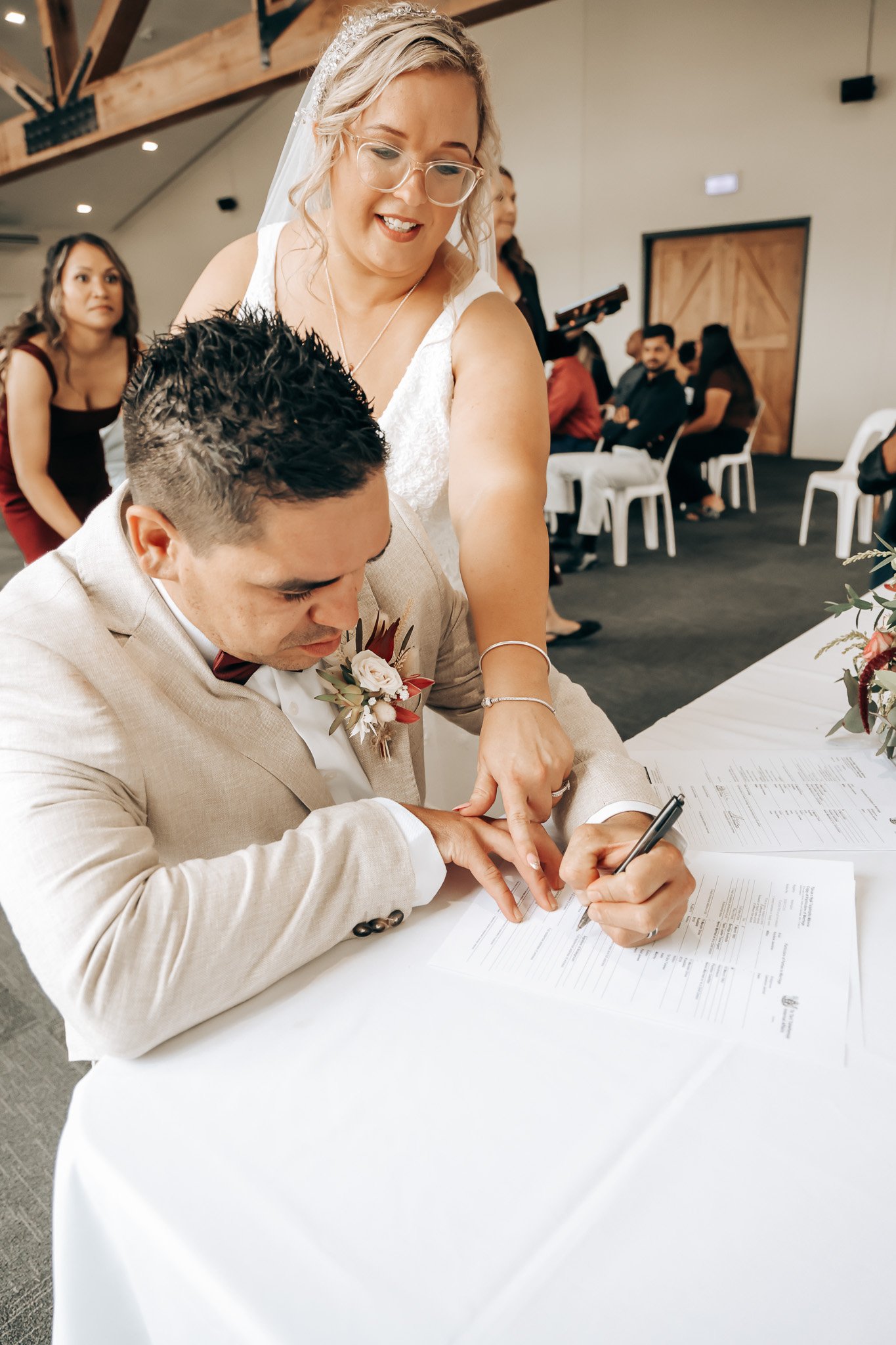 A bride in a white dress and veil standing over a groom signing documents at a wedding ceremony, with guests in the background.
