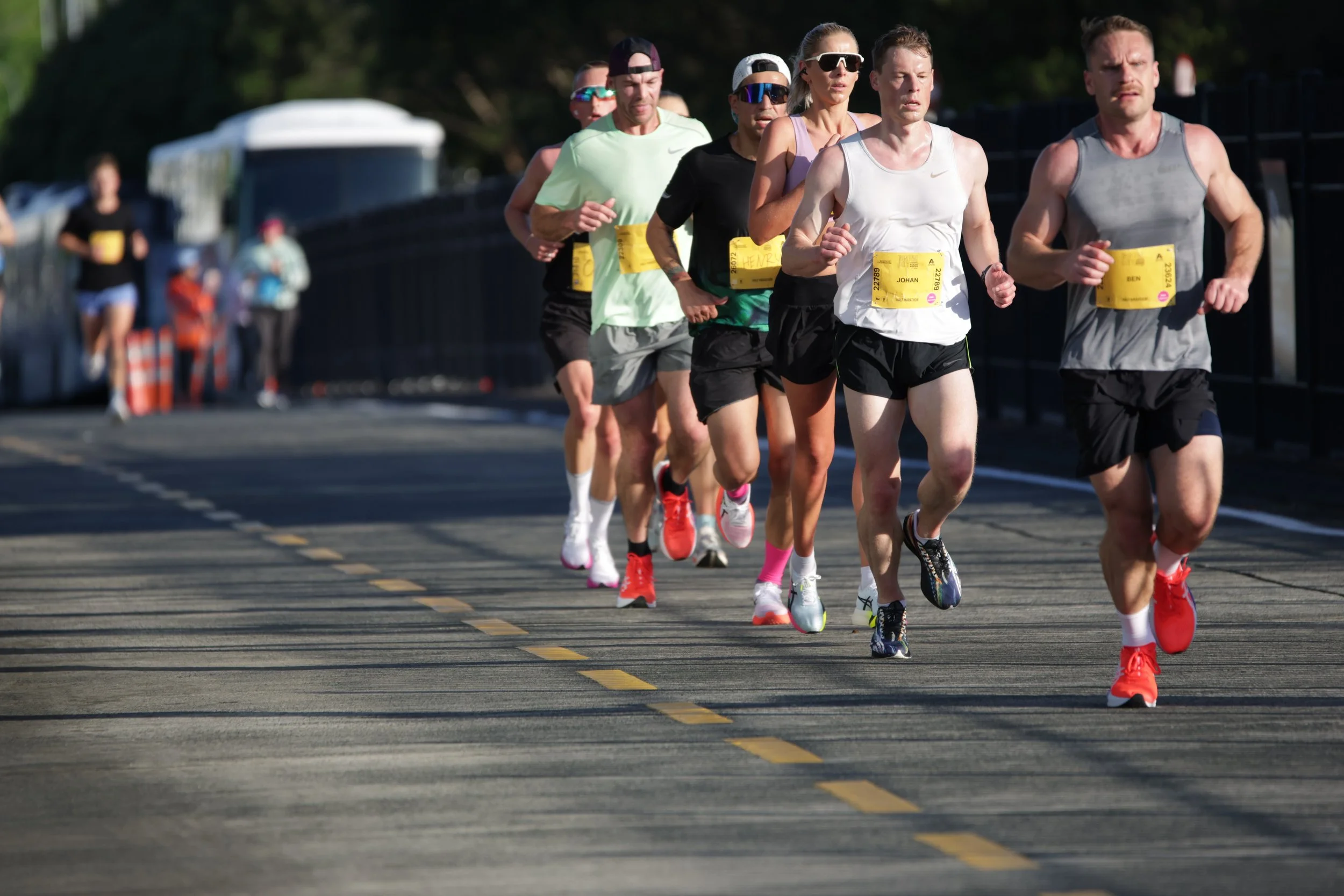 A group of runners participating in a marathon on a paved road with yellow lane markings. The runners are wearing athletic clothing and numbered bibs, with some wearing sunglasses and caps. The background shows blurred spectators and trees.