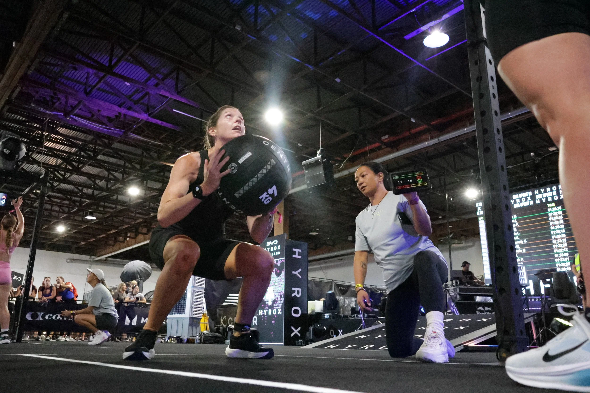 A woman performing a squat with a medicine ball held close to her chest in a gym, with a coach or trainer kneeling nearby, and other people in the background.