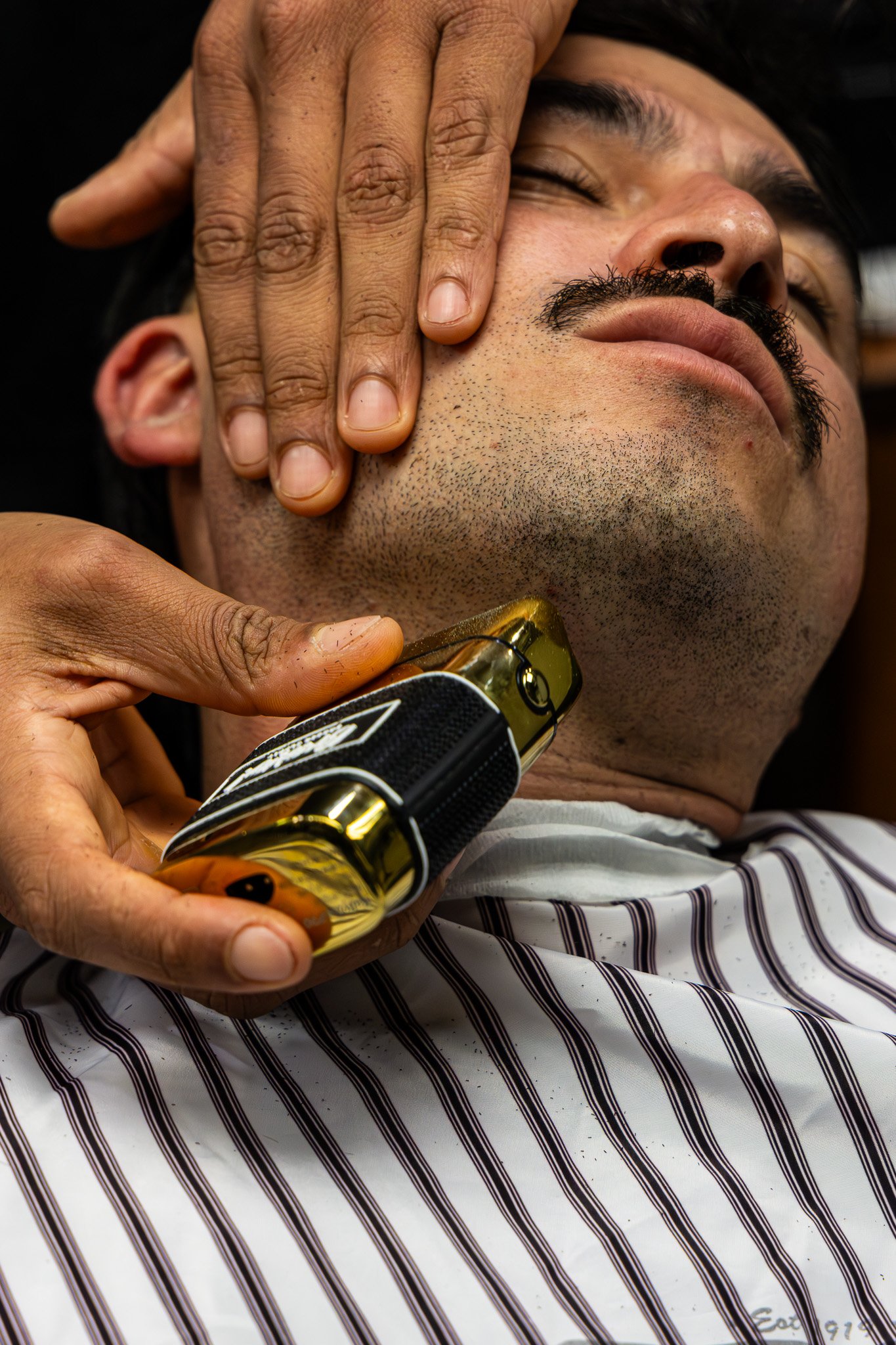 A man with dark hair, a mustache, and stubble is receiving a massage. Someone is using their hand to apply pressure to his face near his cheekbone. The man appears relaxed, with his eyes closed, and is seated wearing a striped shirt. The scene sugges