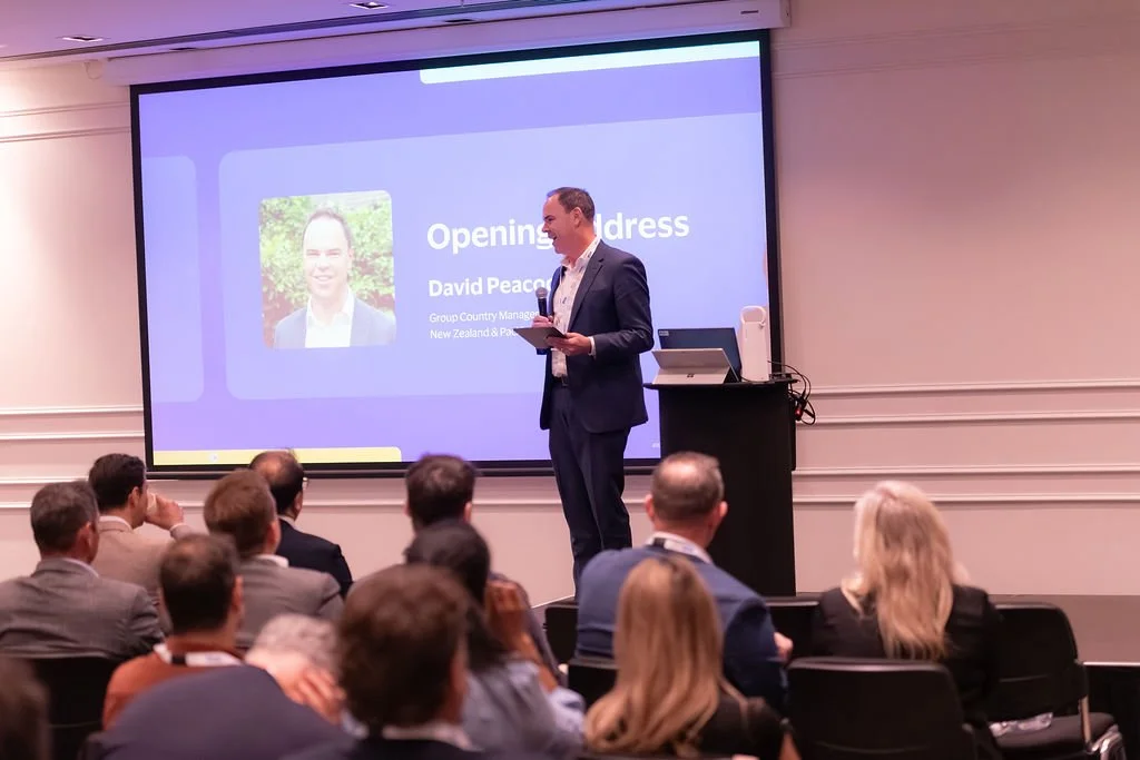 A man in a dark suit giving a speech on stage in front of a large screen at a conference. The screen displays a presentation titled 'Opening Address' with a picture and name of David Peacock, Group Country Manager for New Zealand & Pacific.