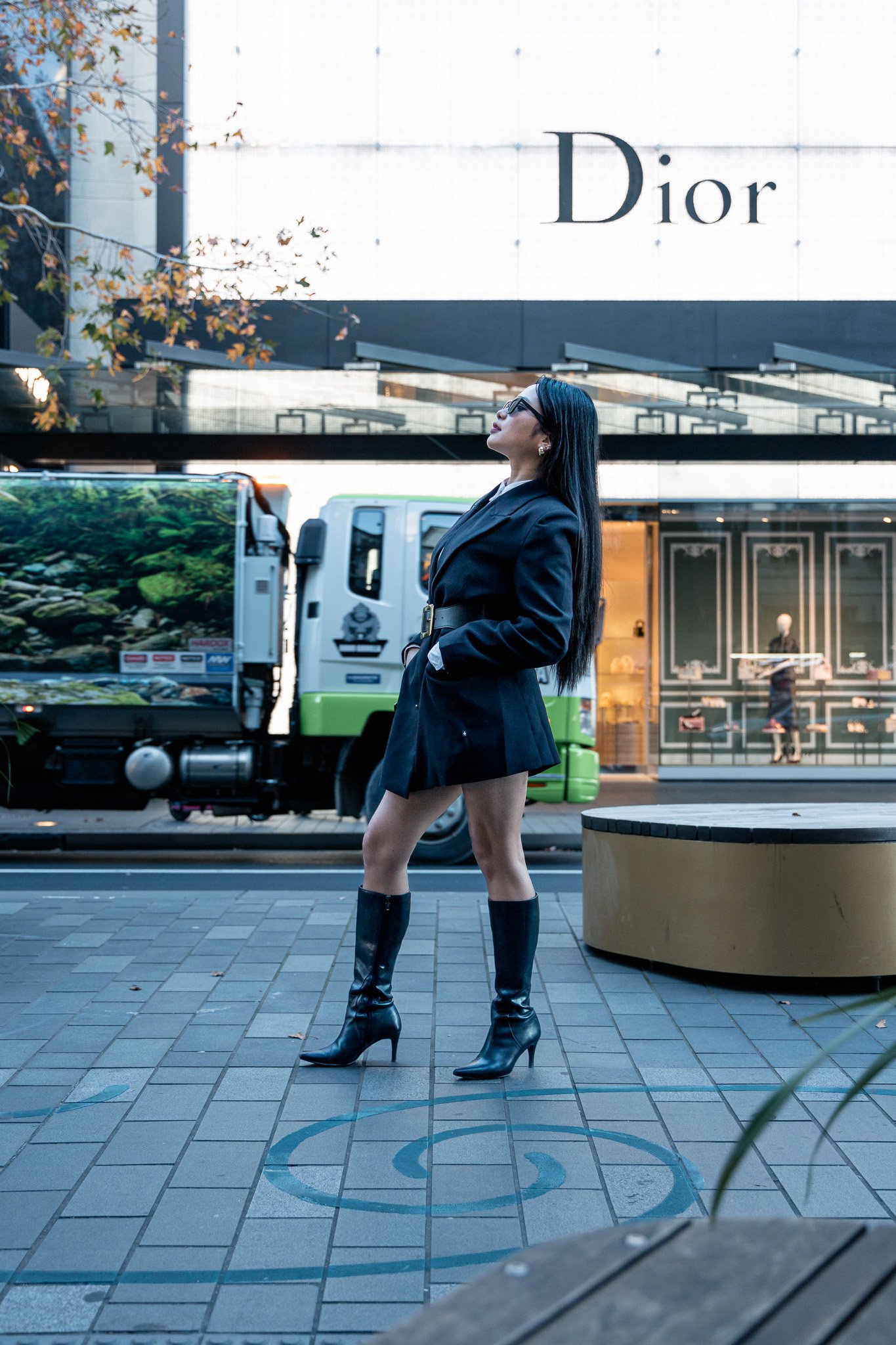 A woman with long black hair, glasses, and a black blazer dress, wearing high-heeled over-the-knee black boots, standing on a city sidewalk in front of a Dior store.