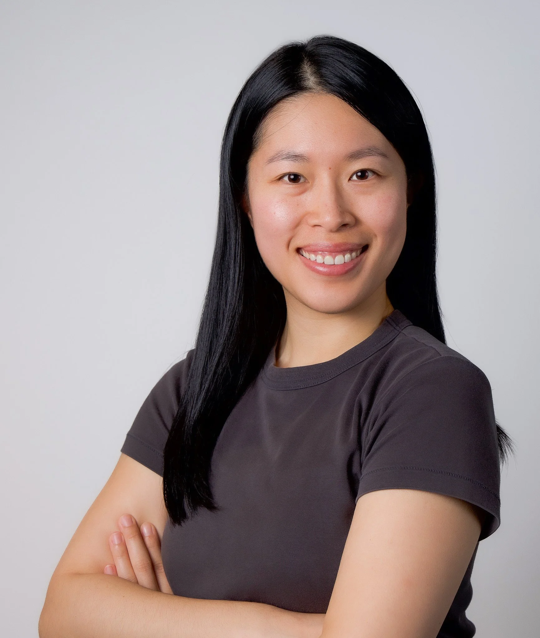 Close-up of a woman with long black hair smiling, wearing a dark t-shirt, posing with crossed arms against a plain white background.