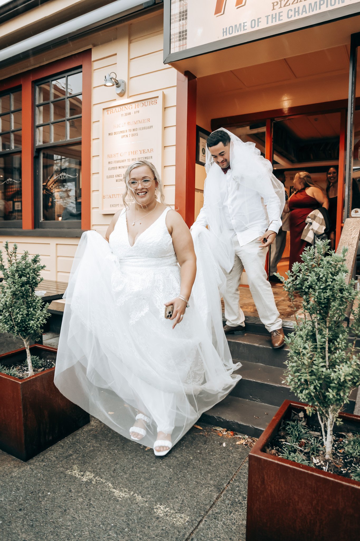 A smiling bride in a white wedding dress and glasses, holding a phone, walking down steps outside a building, followed by a groom in a white shirt and light pants, with women in dresses in the background.