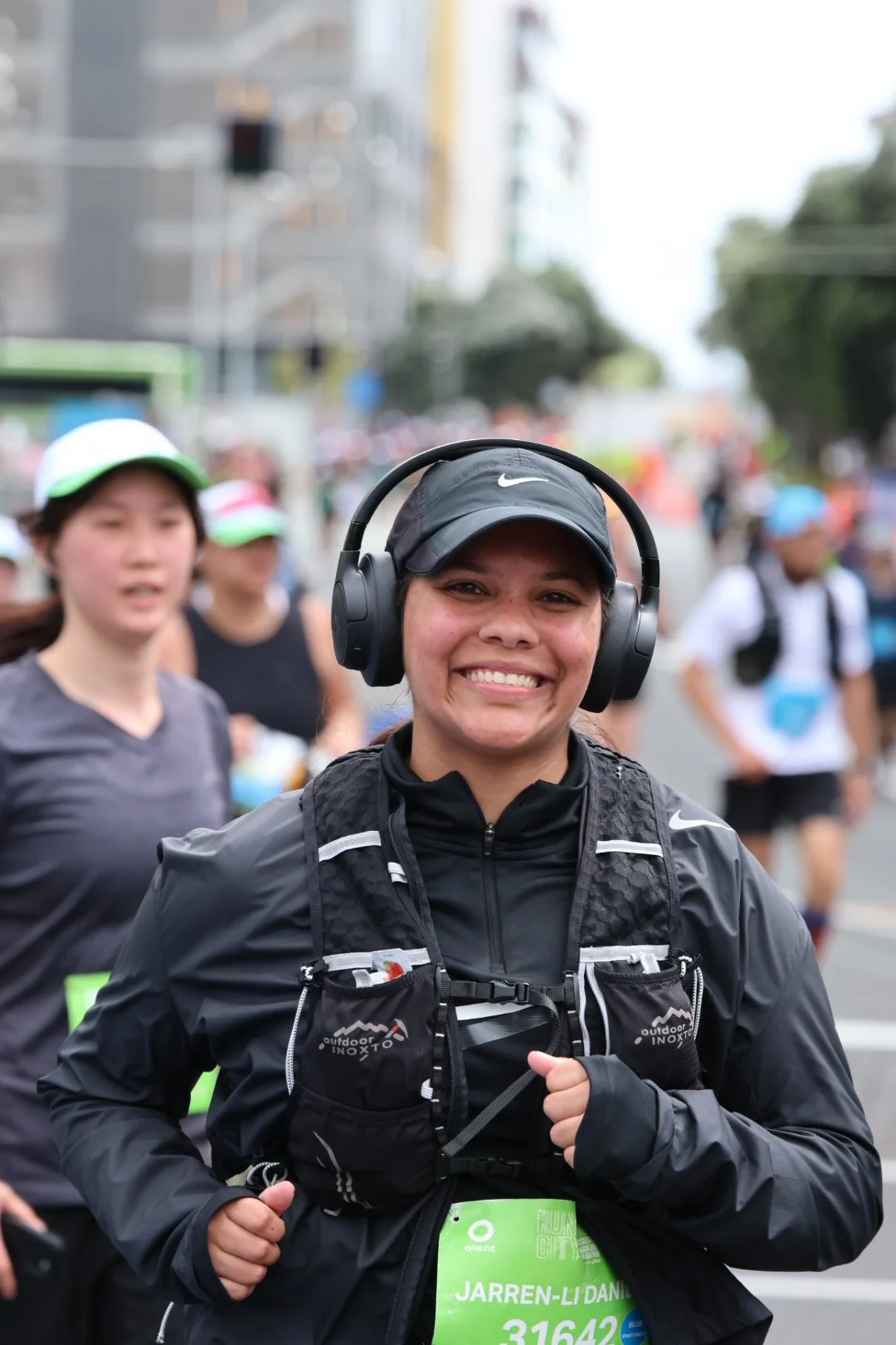 A woman smiling while participating in a marathon, wearing a black Nike cap, headphones, black running gear, and a green bib with her name Jarren-Li Daniel, in a busy urban street with other runners in the background.