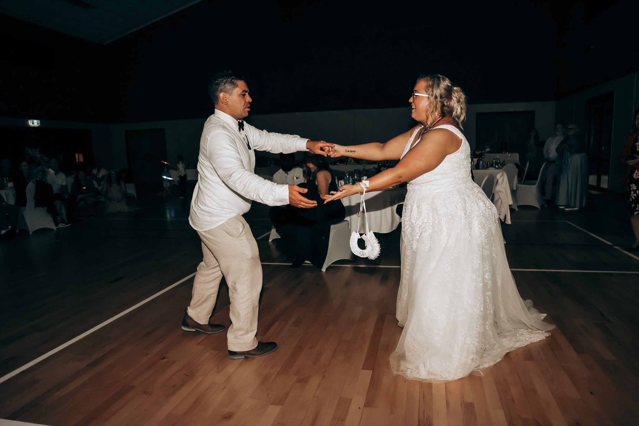 A bride and groom dancing at a wedding reception. The groom, dressed in a white shirt and beige pants, holds the hand of the bride, who is wearing a white wedding gown and glasses, in a dimly lit banquet hall with round tables and other guests in the