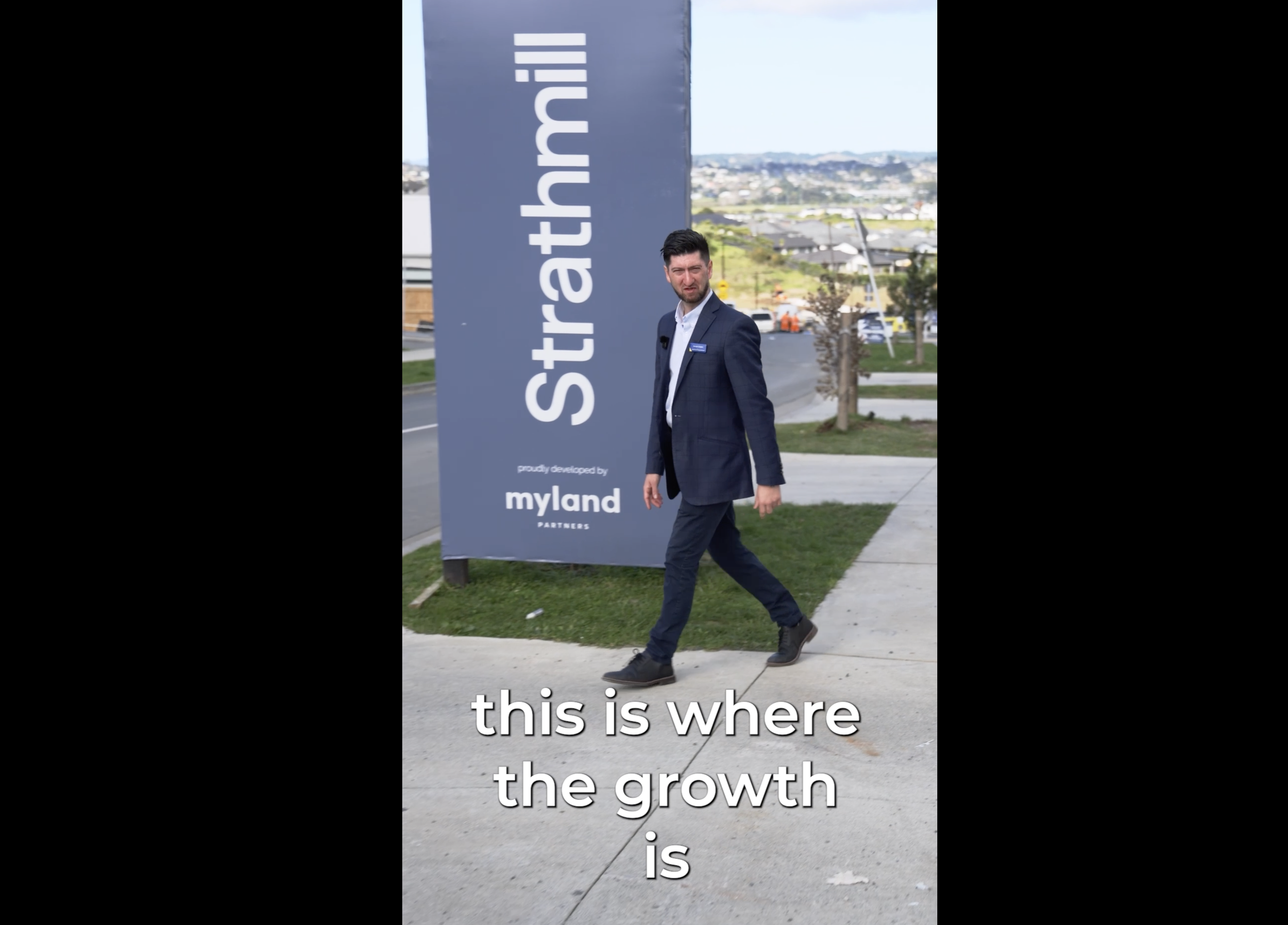 A man in a dark suit walking on a sidewalk next to a large sign that reads 'startabuth'. The background shows a neighborhood with houses, trees, and hills in the distance under a partly cloudy sky.