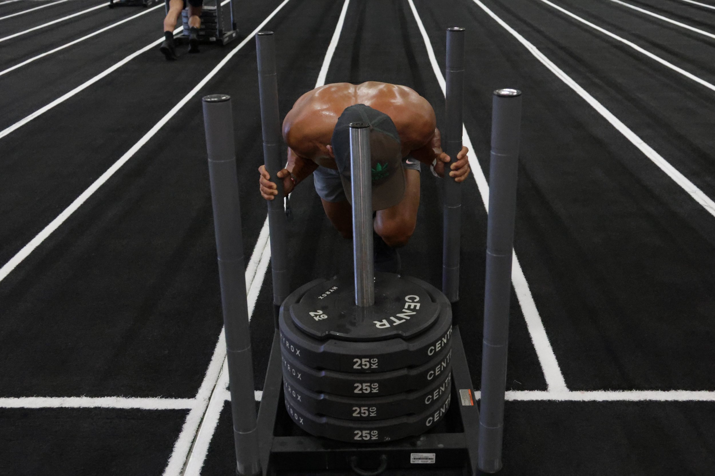 A person bending over on a gym floor, lifting a large weight plate in front of black weight plates stacked on a rack, with running track lines in the background.