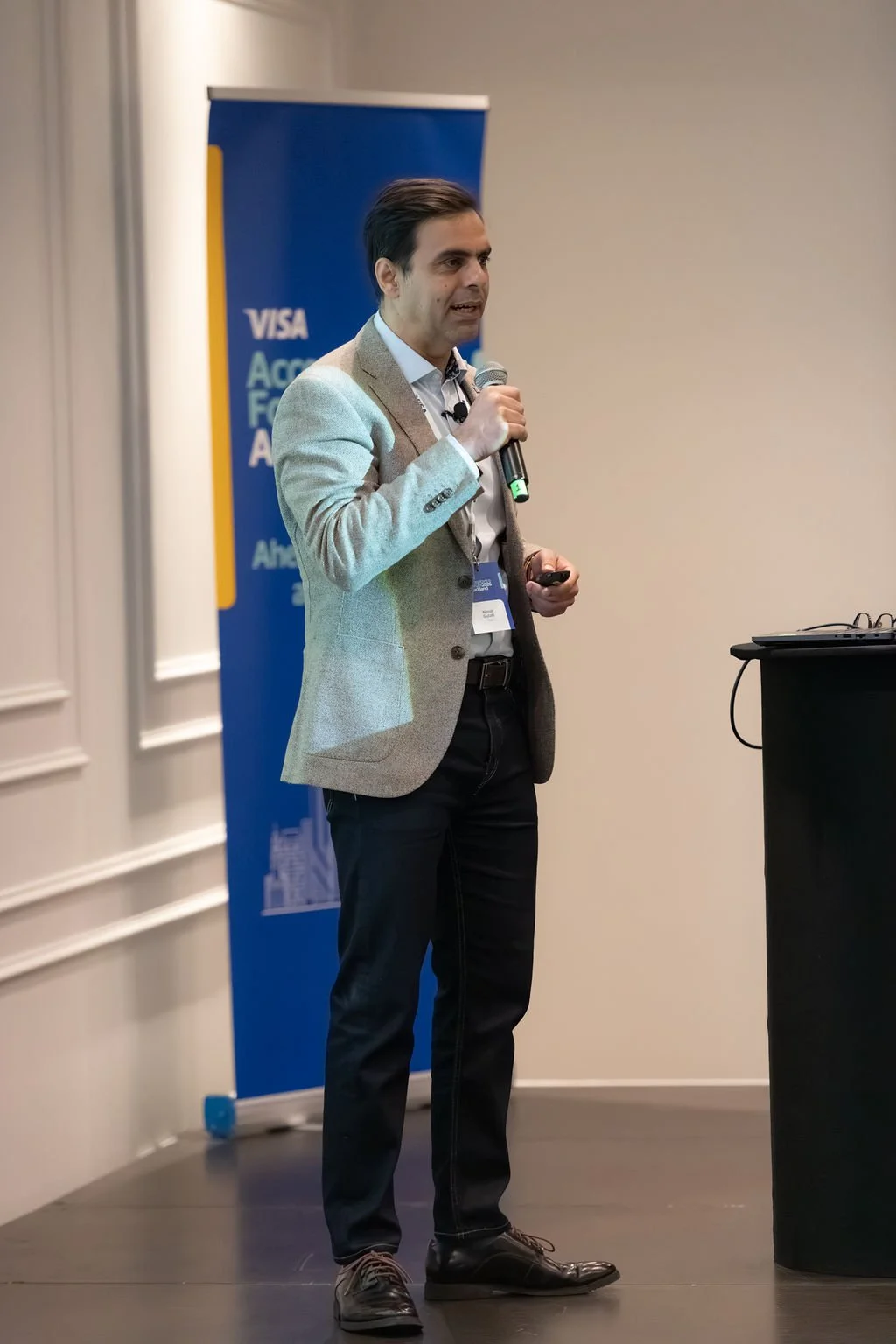 Man in beige blazer giving a presentation with a microphone in front of a blue banner.