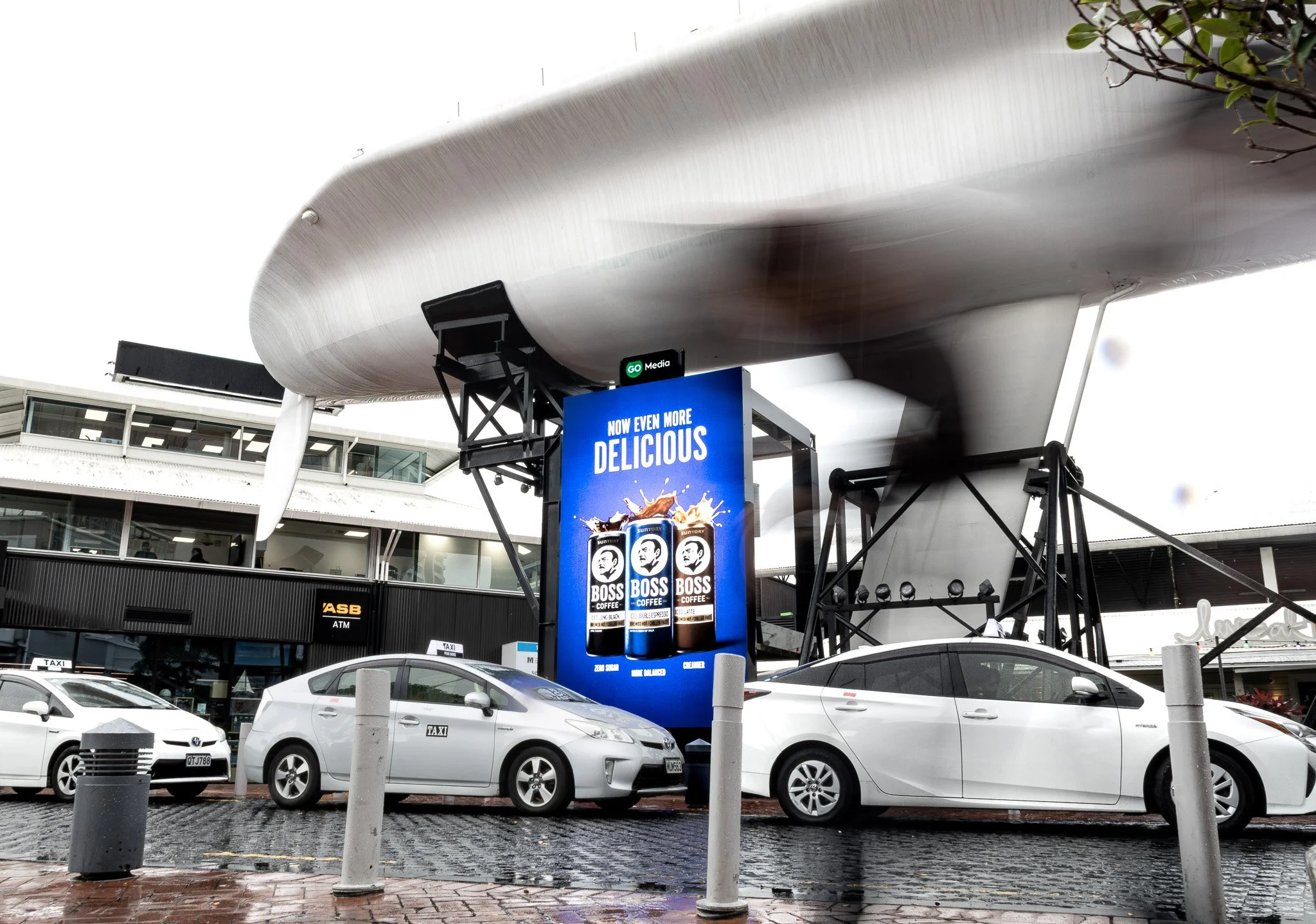 A parking lot in front of a shopping center with several white and gray cars, including taxis, parked along the curb. A large digital billboard displays an advertisement for Boss Coffee, featuring bottles and the slogan "Now Even More Delicious." The