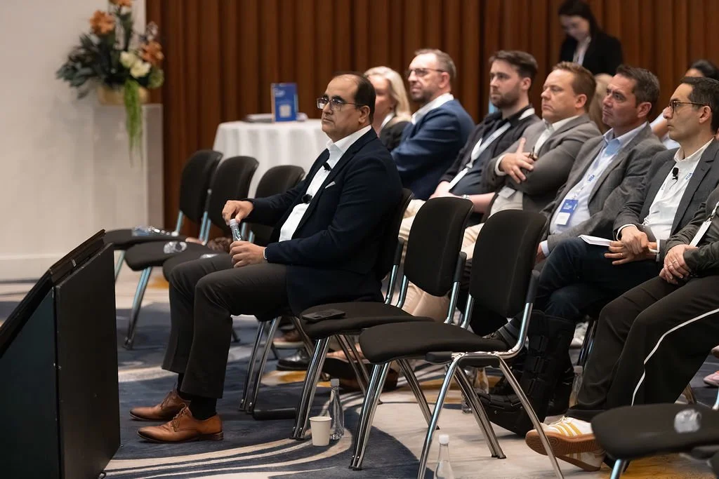 A group of professionals attending a conference, seated in black chairs, listening attentively, with a man in a dark blazer holding a water bottle in the front row.