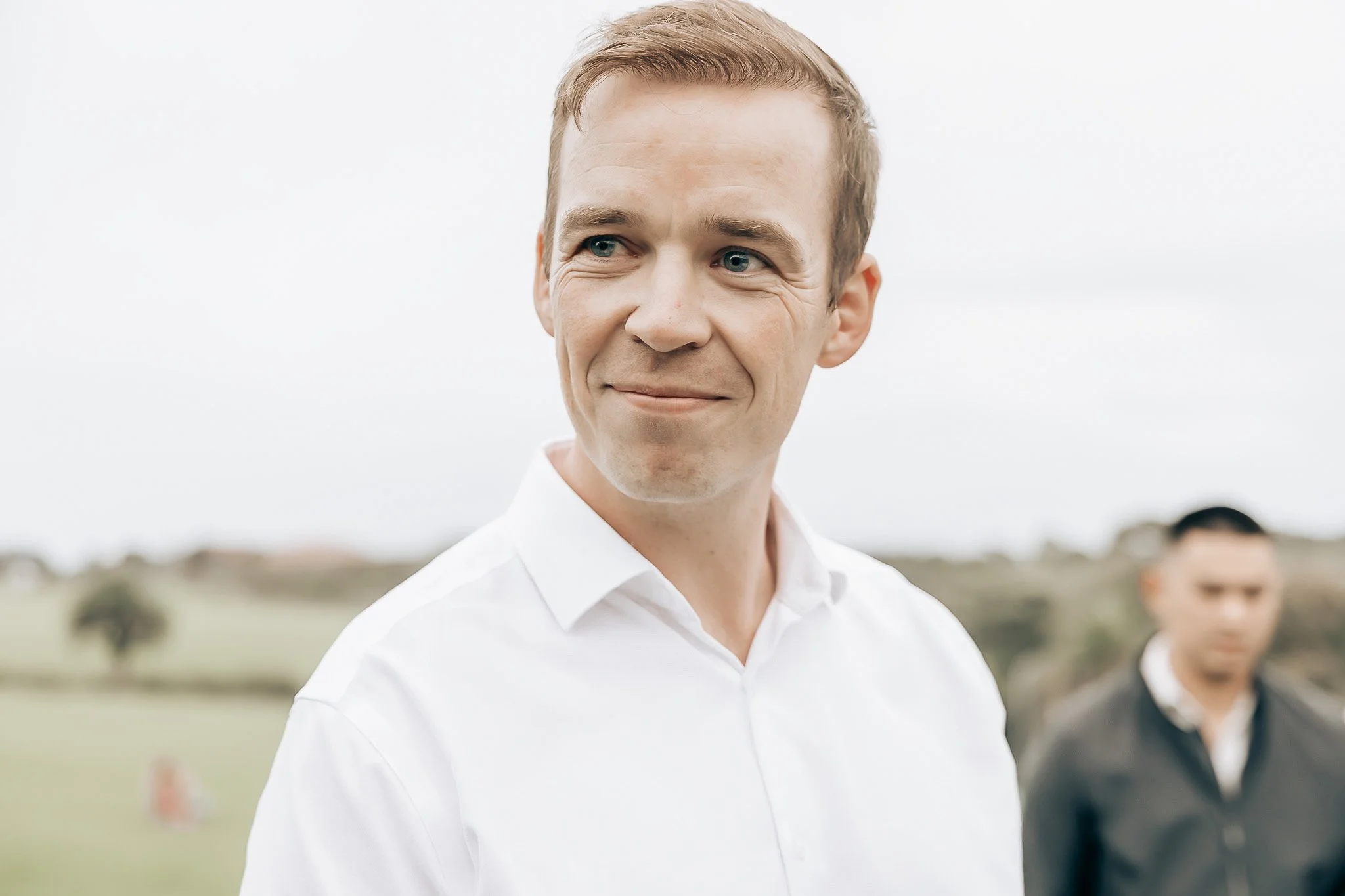 A young man with short, light brown hair, blue eyes, and a slight smile wearing a white collared shirt outdoors on a cloudy day, with blurred greenery and a few people in the background.