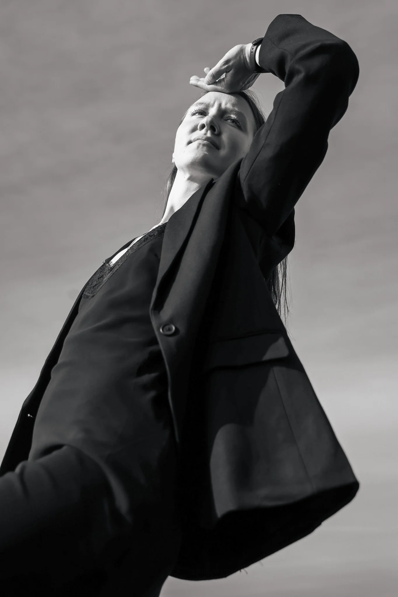 A woman in a suit is standing outdoors, looking up with her hand on her forehead against a cloudy sky.