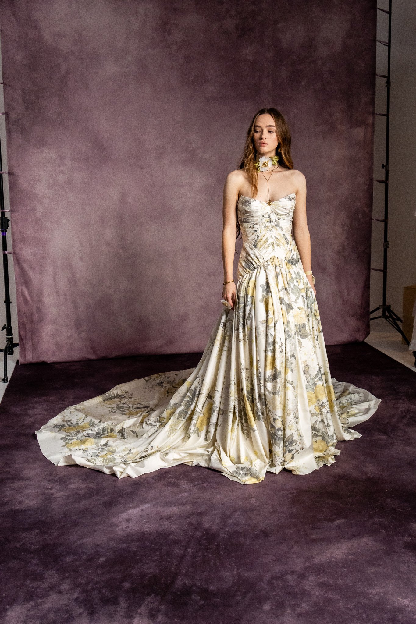 A woman wearing a strapless, floral-patterned gown with a long train standing against a purple backdrop.