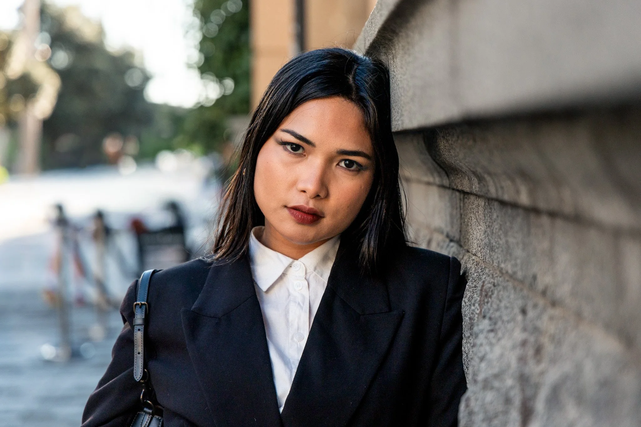 A woman with long black hair, wearing a white blouse and black blazer, standing against a brick wall on an outdoor street.