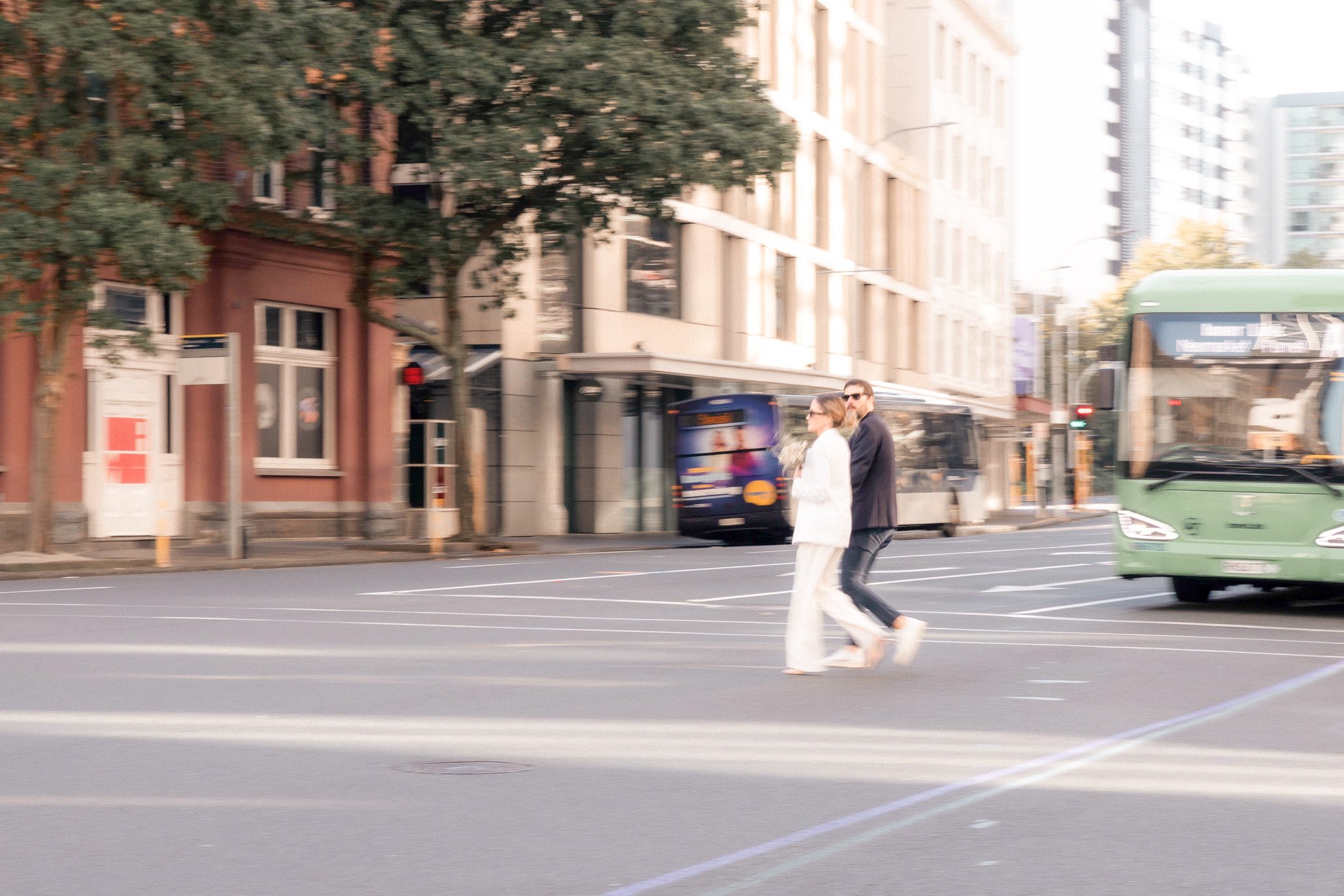 Two people, a man and a woman, crossing a city street at a crosswalk, with buildings, trees, and a green bus in the background.