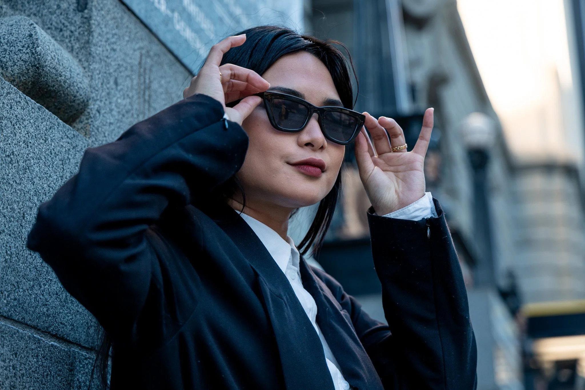 Woman with dark hair and lipstick adjusts black sunglasses while sitting on a stone bench outdoors.