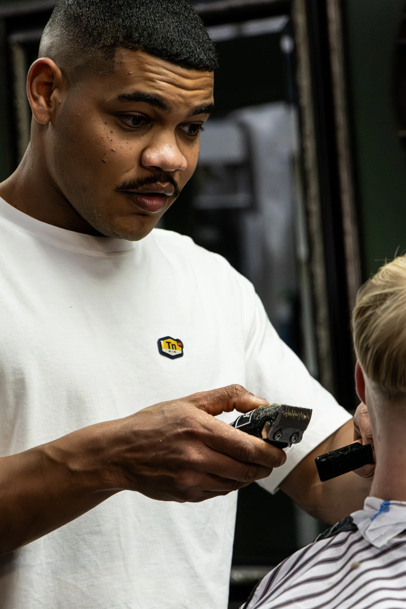 A man with a short haircut and mustache is holding a black and silver hair clipper and showing it to a young boy with light-colored hair. The man is wearing a white T-shirt with a small black and yellow logo.