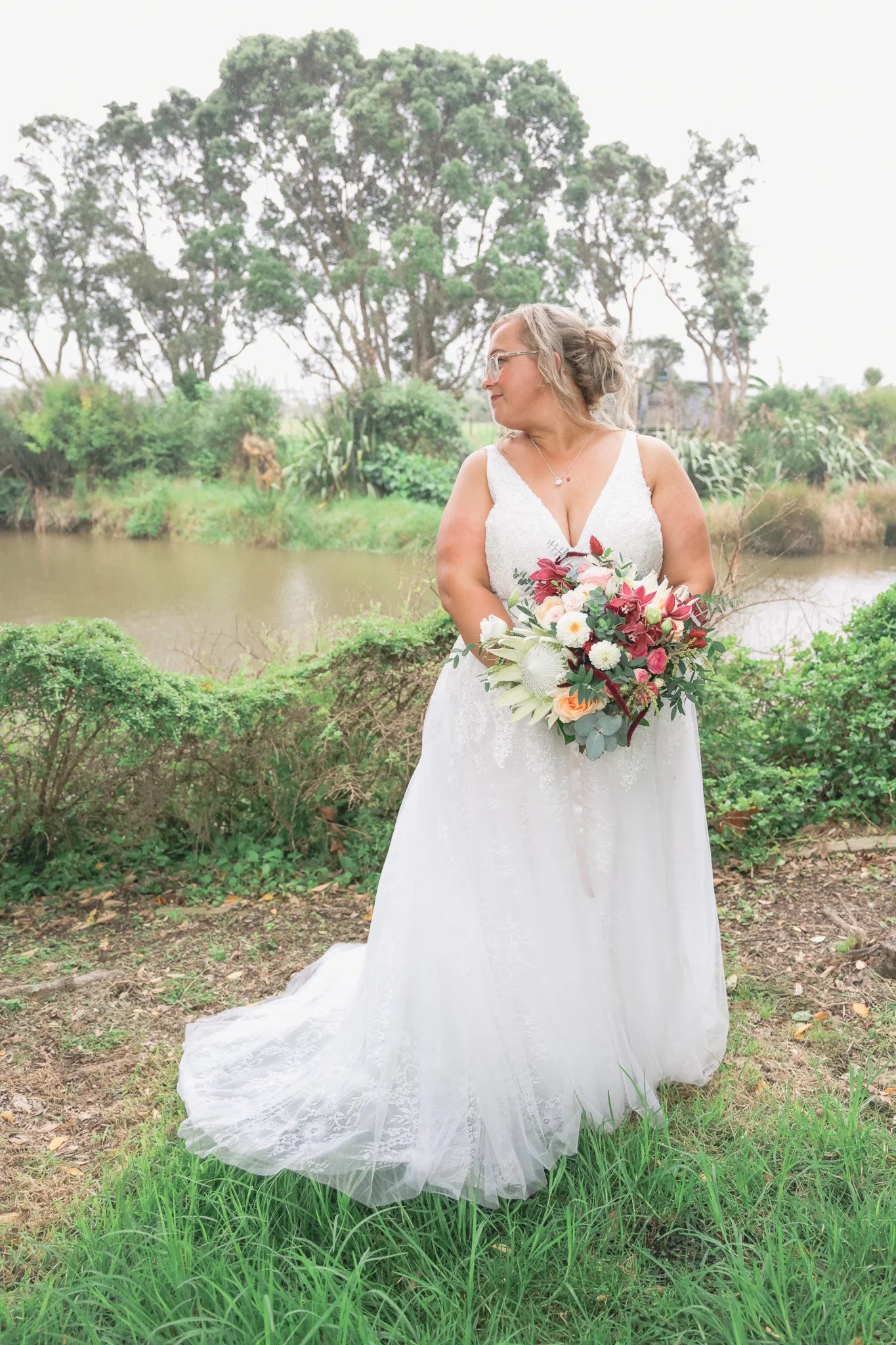 A woman in a white wedding dress holding a bouquet of flowers stands outdoors near a pond, with trees and green shrubbery in the background.