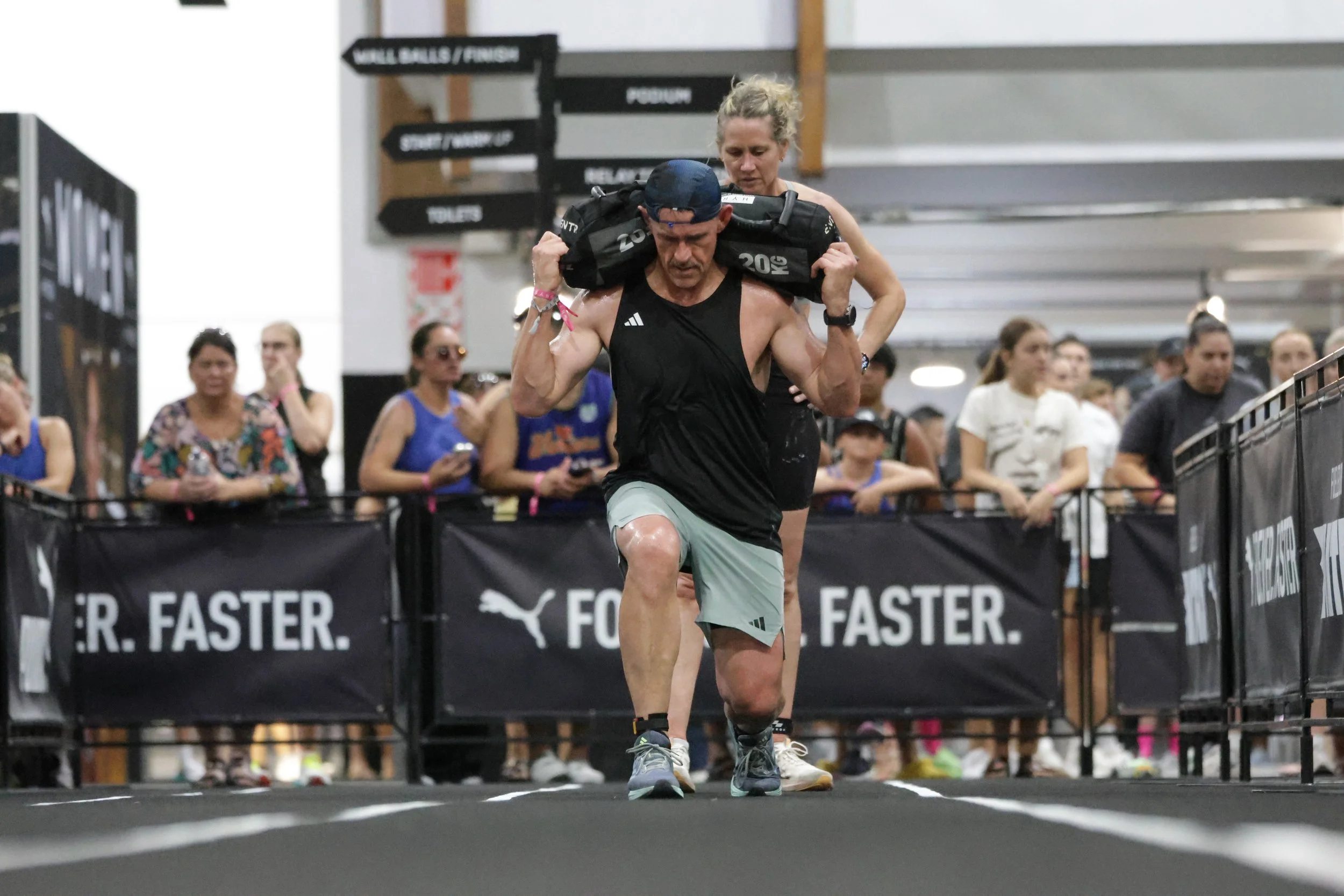 A man running in a race carrying a weighted sandbag on his shoulders with a woman running closely behind at an indoor event. Several spectators are in the background watching the race.