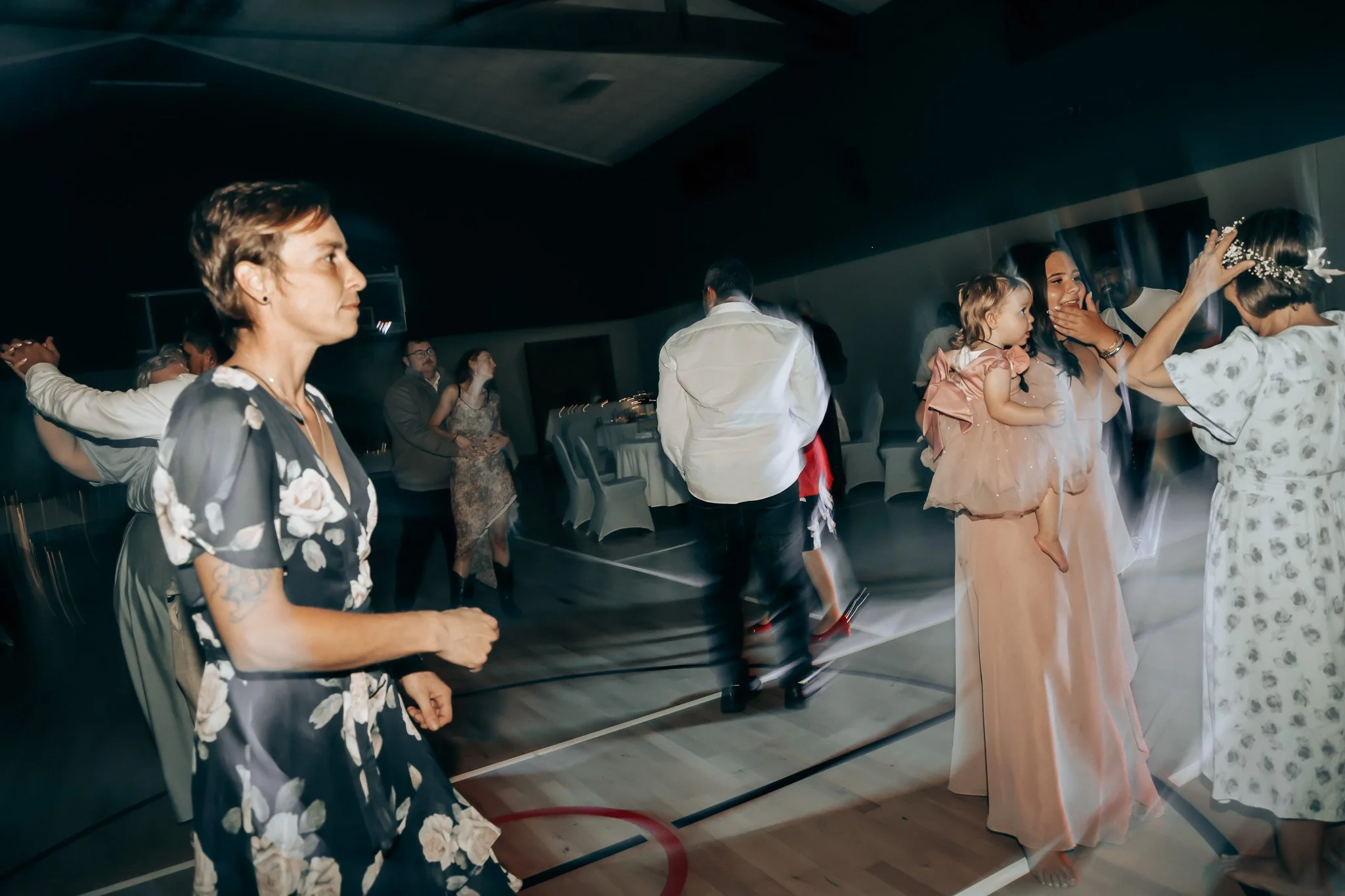 People dancing at a celebration, including a woman with a blonde bun and a floral wedding dress, and a little girl in a pink dress, with other guests in the background.