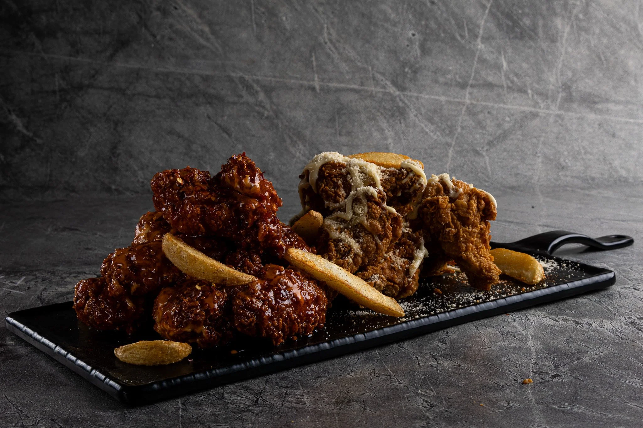 Fried chicken pieces topped with barbecue sauce and potato chips on a black rectangular platter, garnished with grated cheese and served with a drizzled white sauce on the side, set against a dark textured background.