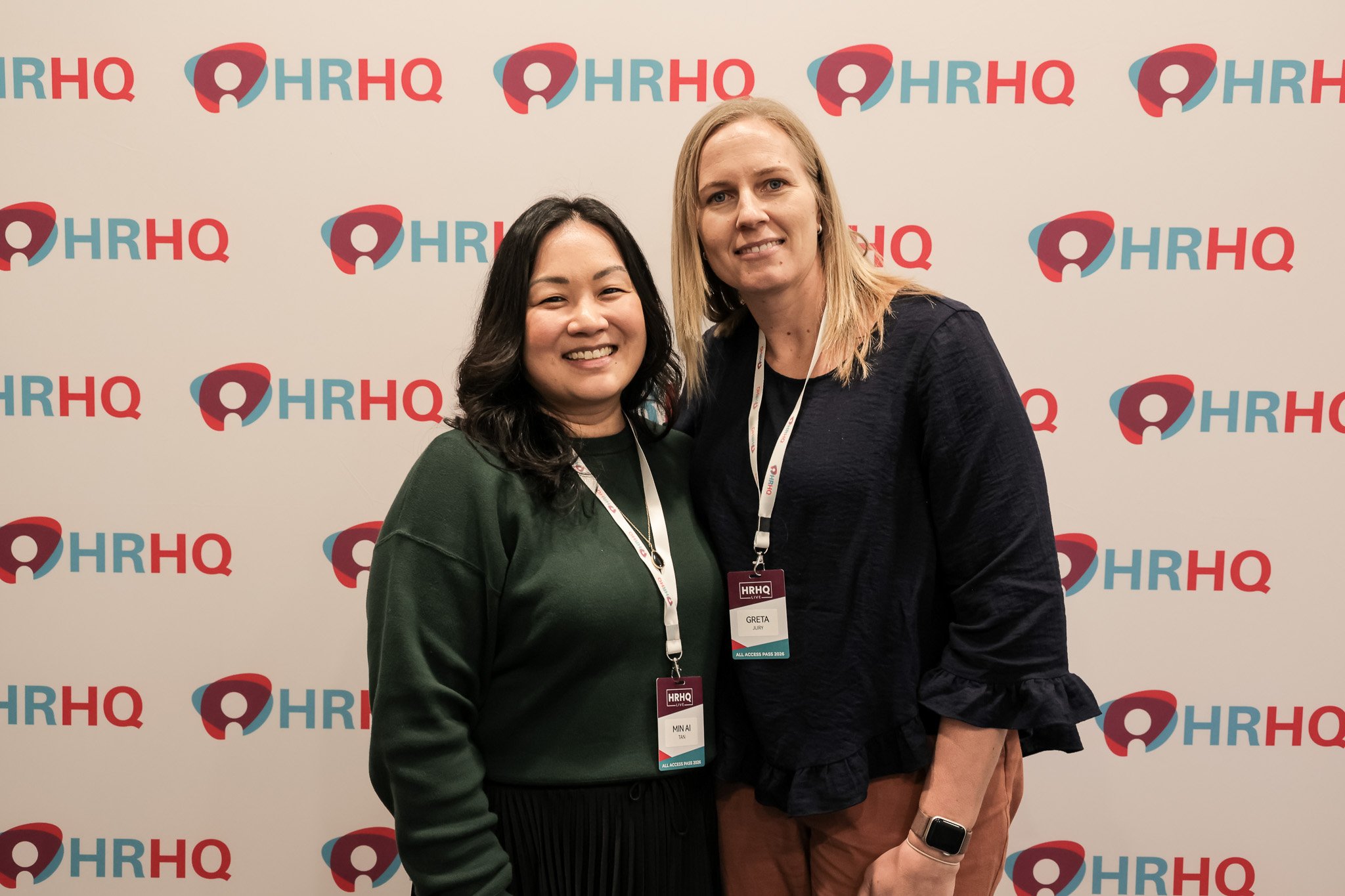 Two women standing together at a conference, smiling, with a background featuring the HRHQ logo repeatedly. One woman has dark hair and is wearing a dark green top; the other has blond hair and is wearing a dark top, both with conference badges aroun