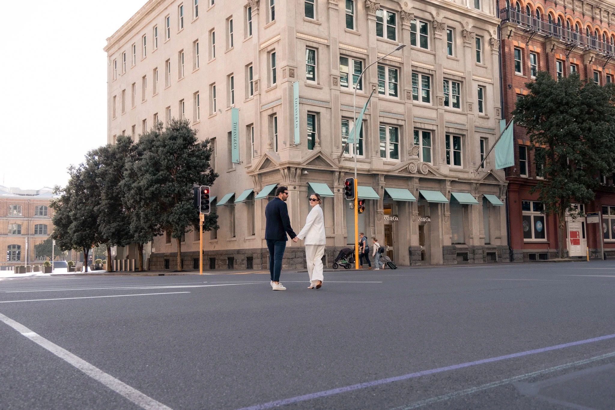 Two people holding hands and walking across a city street in front of a large building with Tiffany & Co. banners, trees, and traffic lights.