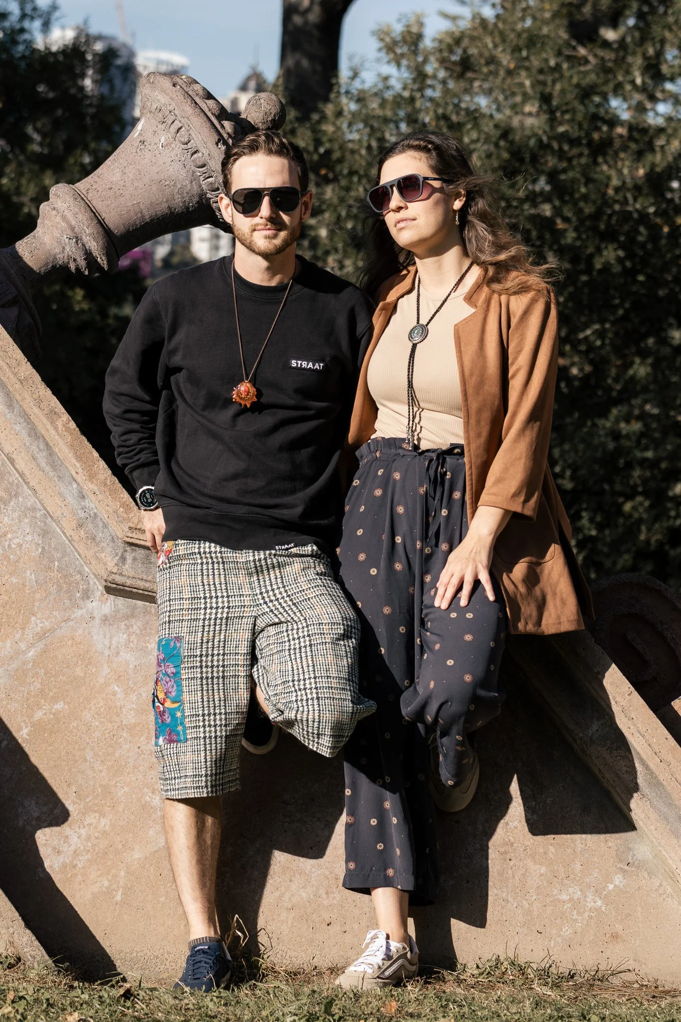 Two young adults, a man and a woman, standing outdoors on a sunny day near a large sculptural object with trees in the background. They are both wearing sunglasses and casual clothing, with the woman wearing a beige top and brown jacket, and the man 