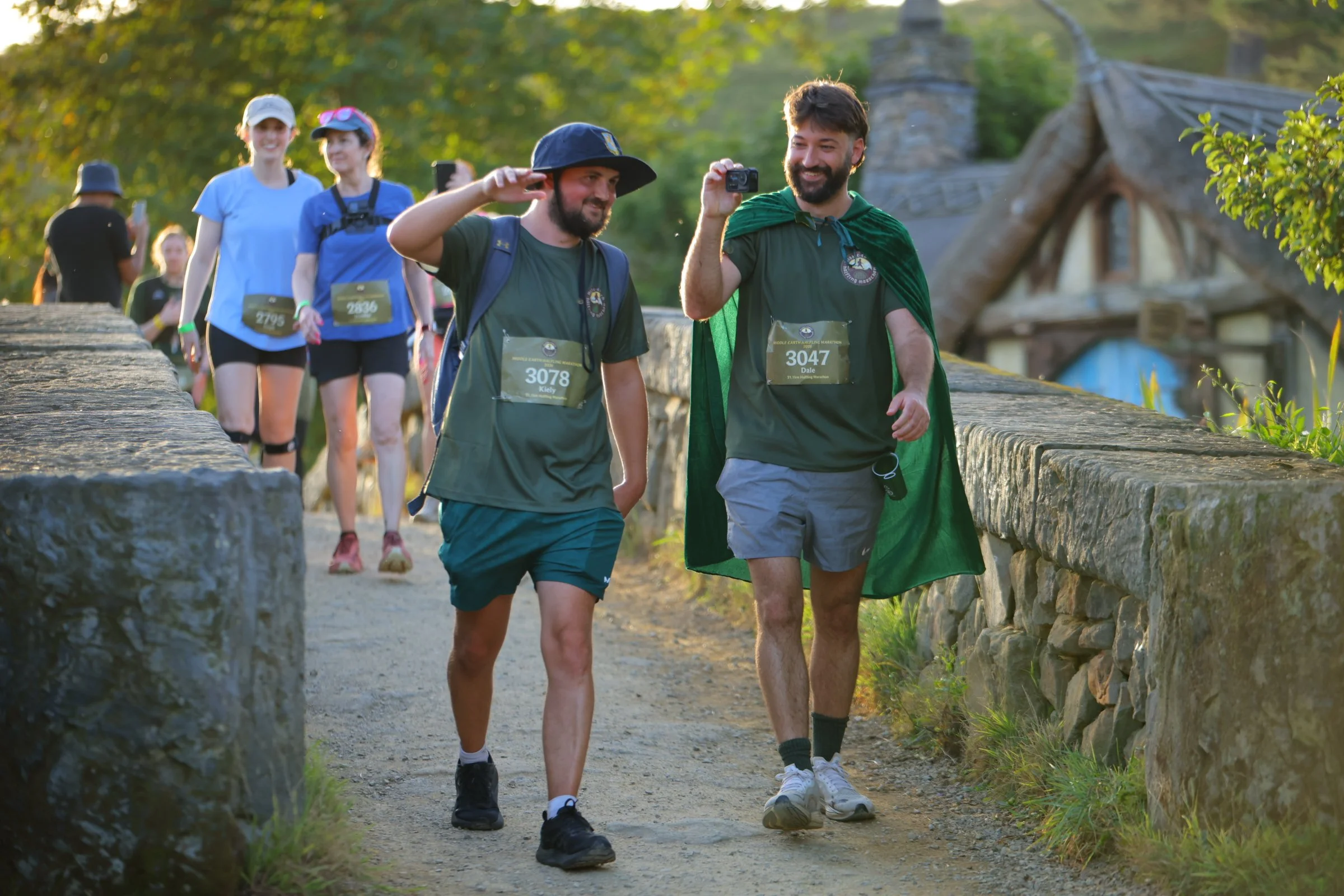 Group of runners participating in a marathon, walking along a stone bridge with lush green trees in the background, some taking photos.