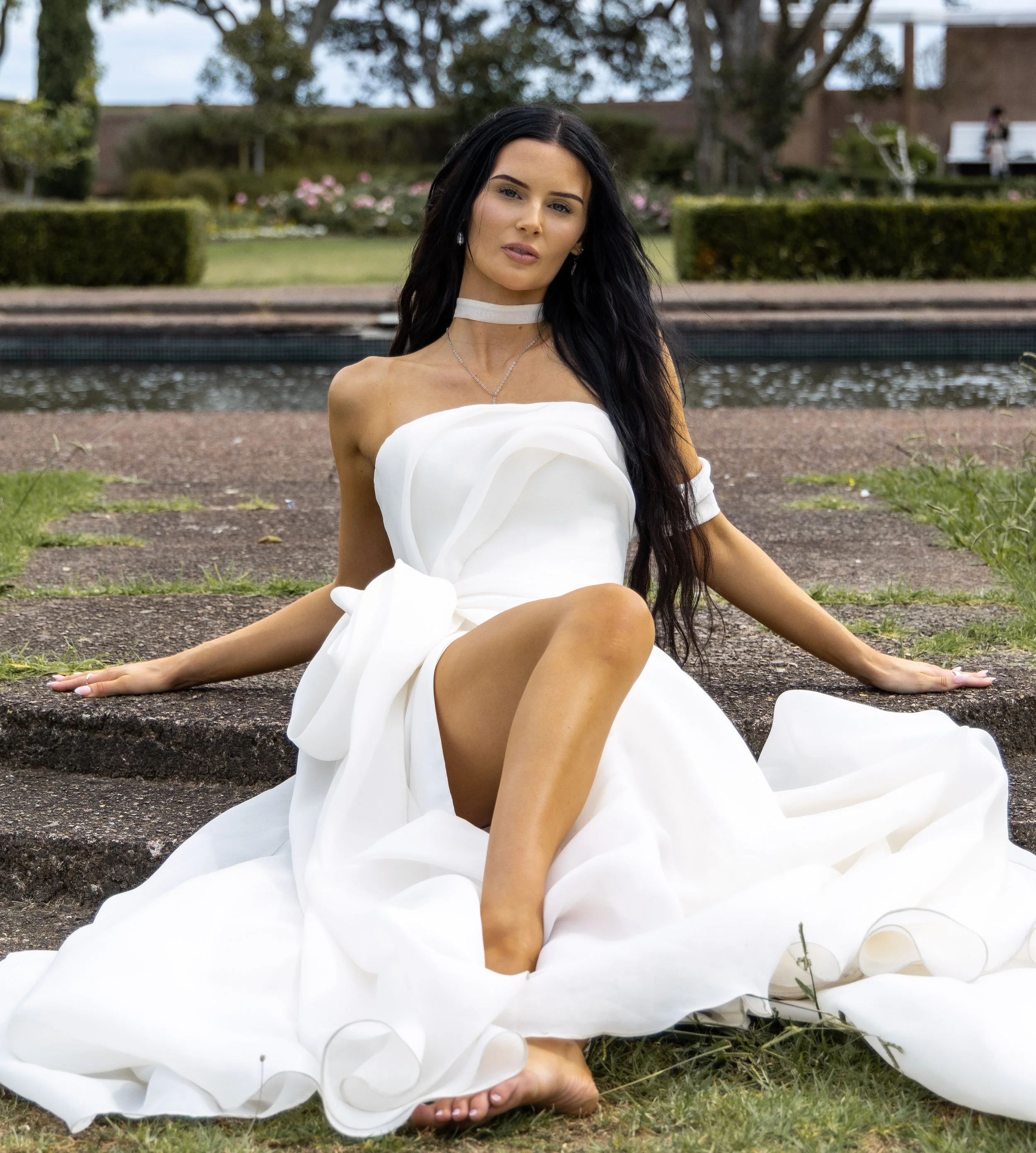 A woman in a white dress sitting on the ground outdoors with a pond and trees behind her.