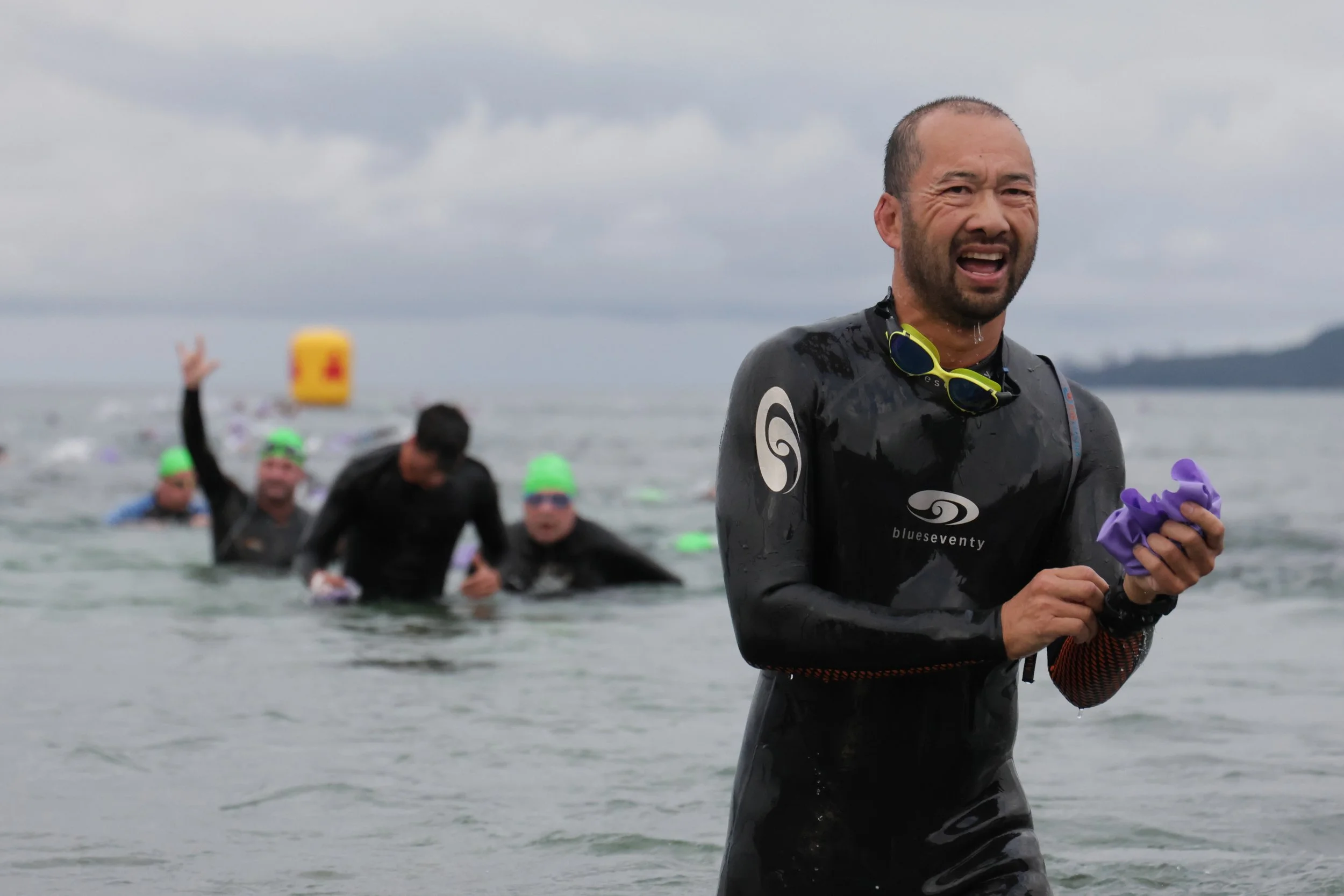 A man in a wetsuit holding purple goggles and smiling on the beach during a swimming event with other swimmers in the background